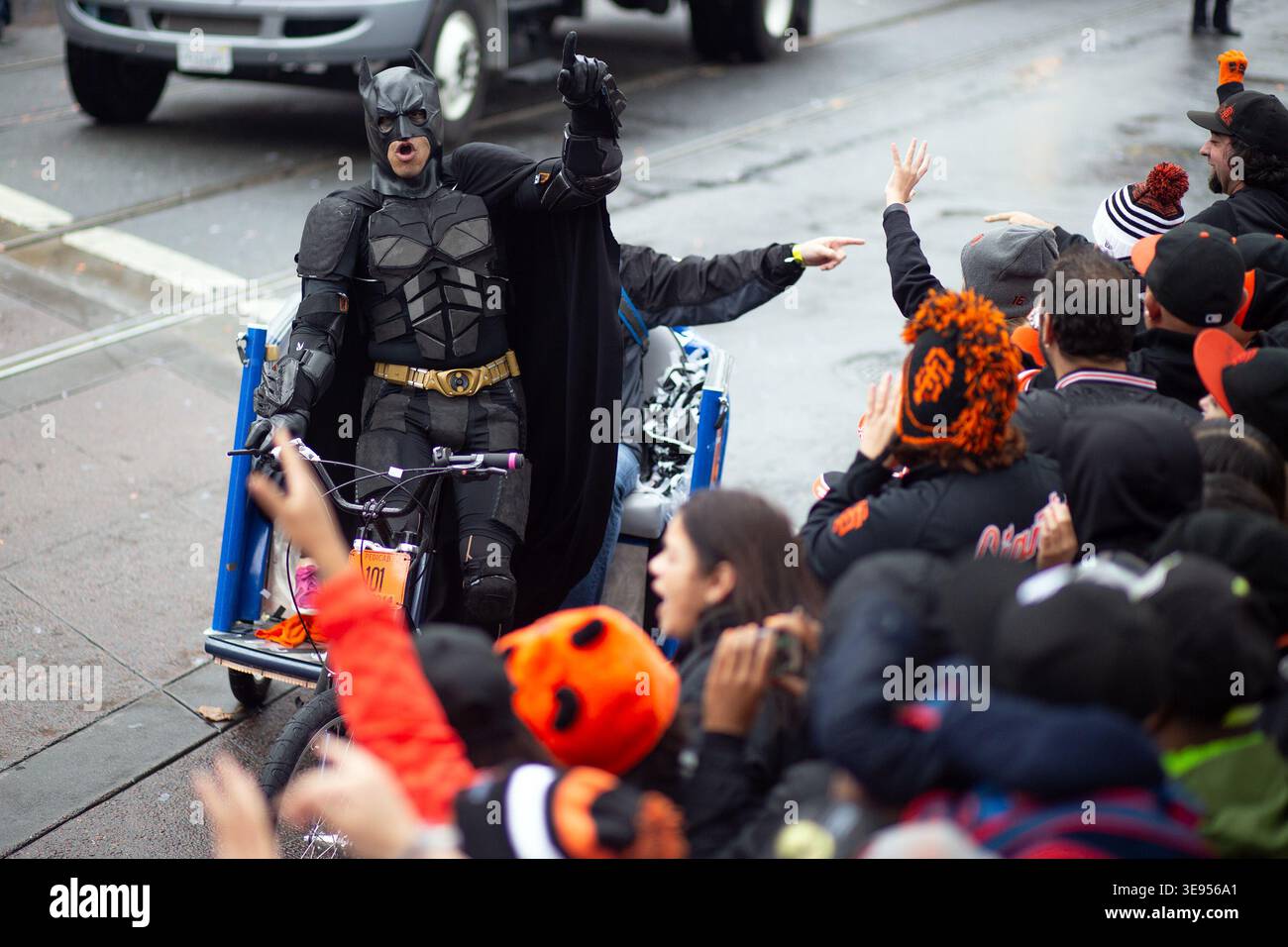Una persona in costume di Batman cavalca un triciclo attraverso una folla di persone. Gli spettatori sventolano e applaudono mentre il personaggio passa. Foto Stock