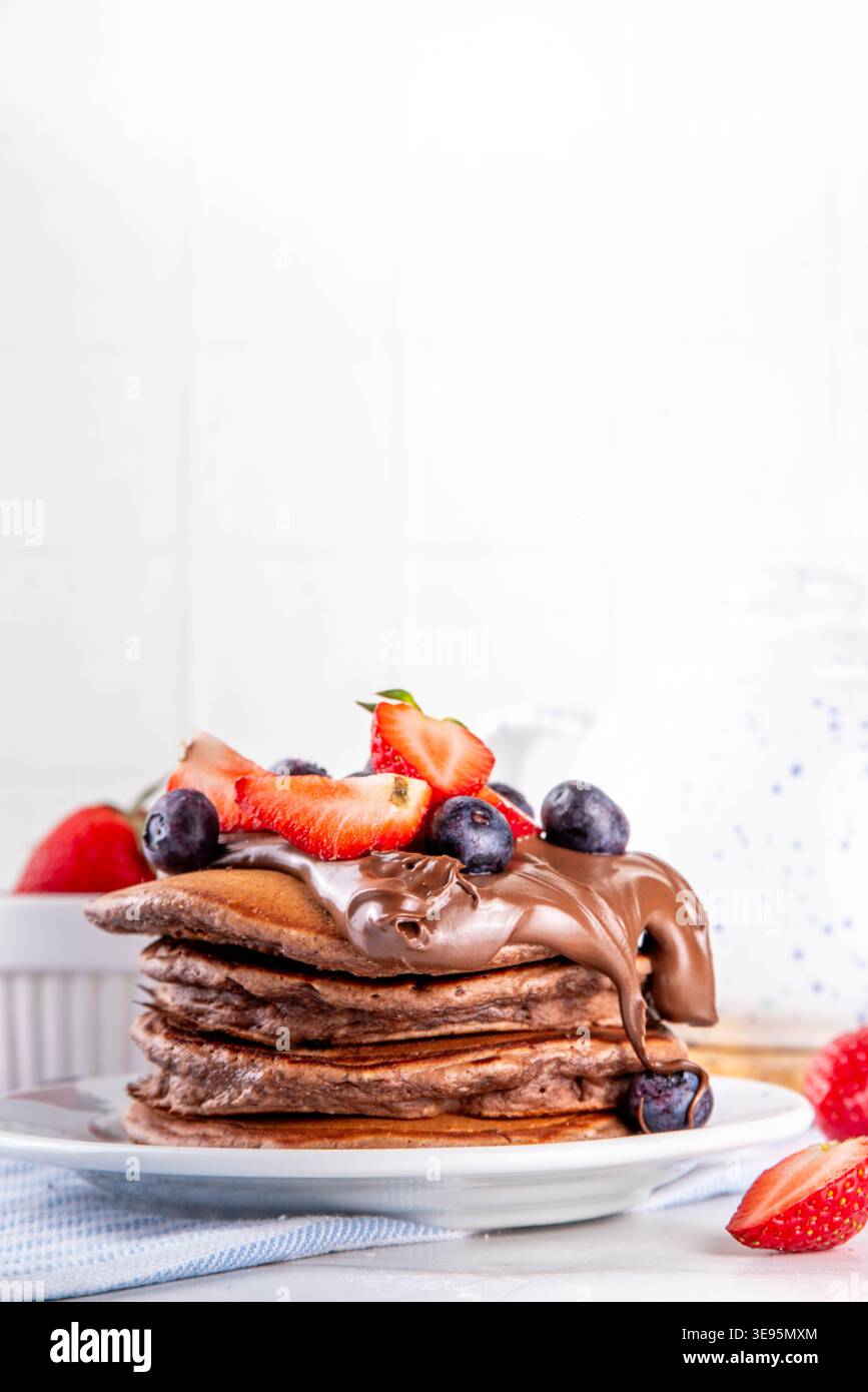 Pila di pancake al cioccolato con una ricca crema di pasta al cioccolato e frutti di bosco freschi - fragole, mirtilli. Colazione dolce e gustosa Foto Stock