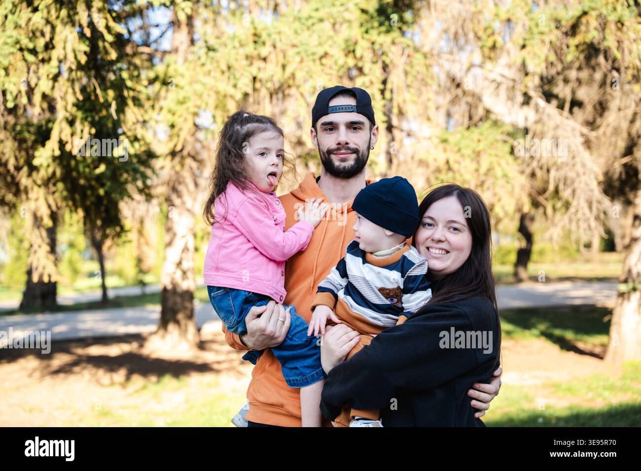 Famiglia con padre, madre e due bambini in piedi nel parco che tengono i bambini e guardano la macchina fotografica con espressioni calde. Concetto di legame familiare e pa Foto Stock