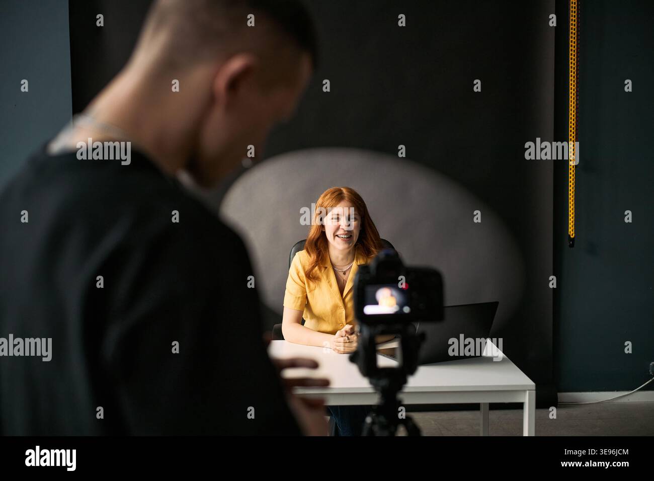 Ripresa media di una giovane donna caucasica che ospita lo streaming online, che parla con la fotocamera mentre un videografo filma i contenuti alla scrivania. Ideale per l'economia dei creator, il blog e l'uso del marketing digitale Foto Stock