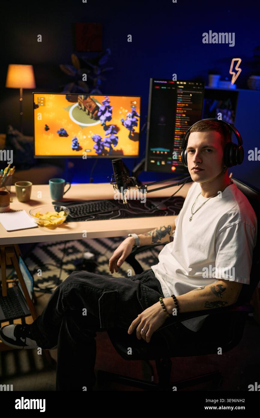 Ritratto di un giovane uomo caucasico che indossa le cuffie, streaming di contenuti di videogiochi con configurazione a doppio monitor e microfono. Ideale per canali di gaming, economia dei creatori, marketing broadcast live, verticale Foto Stock