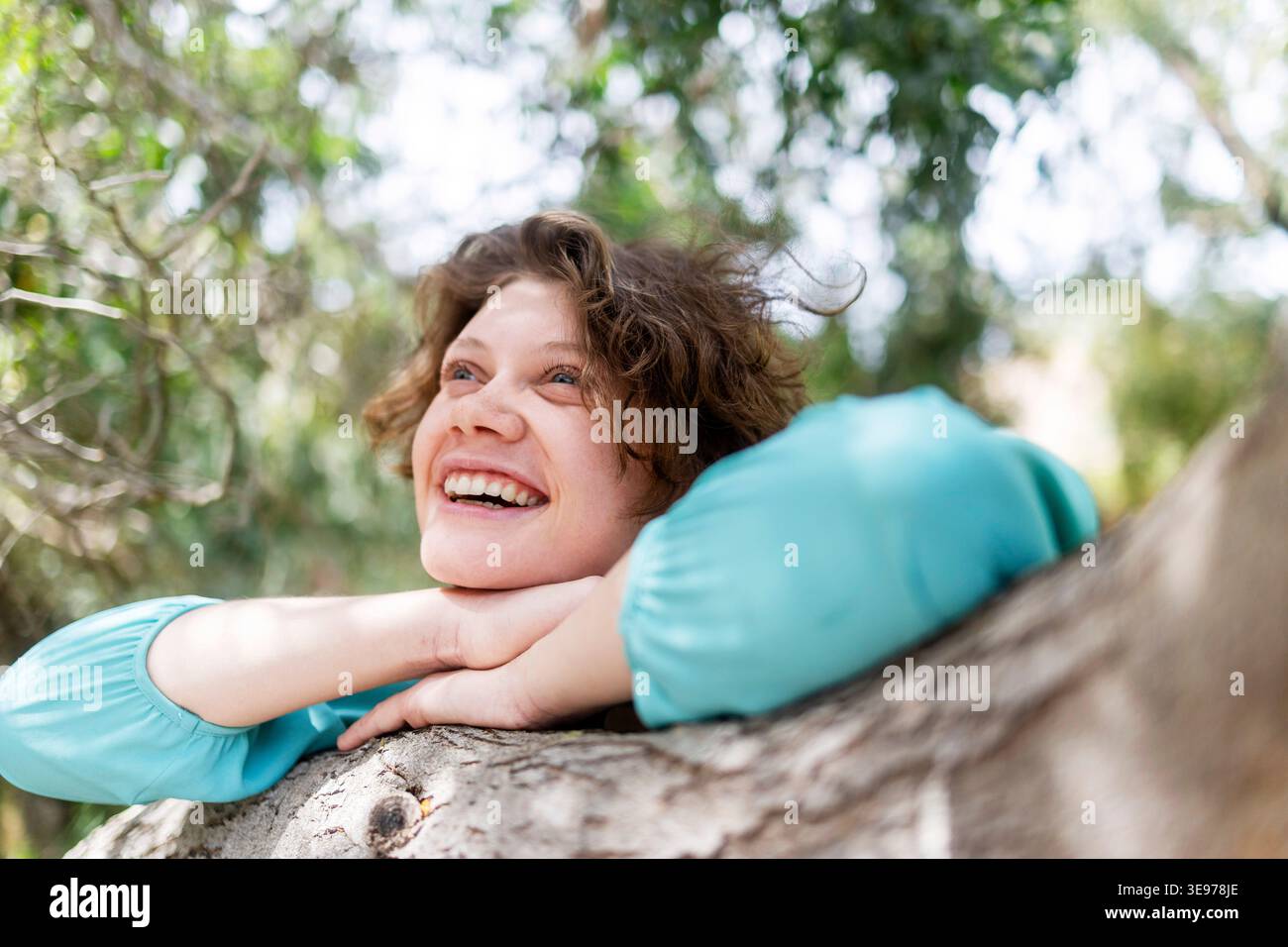 Un ritratto ravvicinato di una donna che ride con ricci e lentiggini disordinati che appoggiano il mento su un tronco di albero. Foto Stock