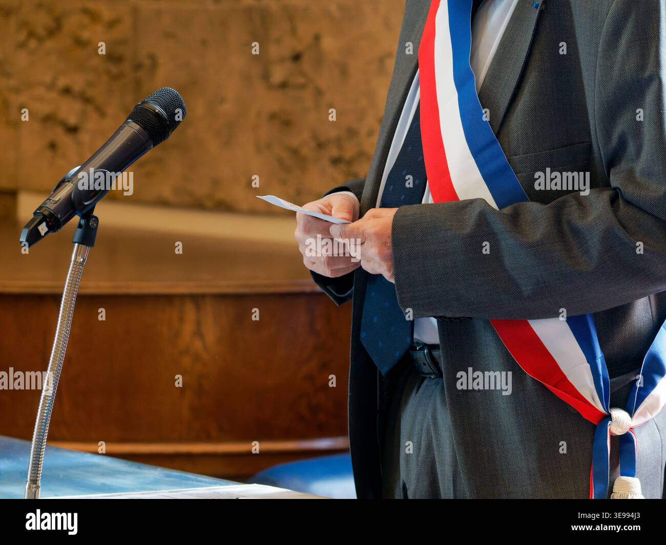 Primo piano di un sindaco francese che indossa una fascia tricolore durante una cerimonia ufficiale, in attesa di documenti su una scrivania in un ambiente civico. Foto Stock
