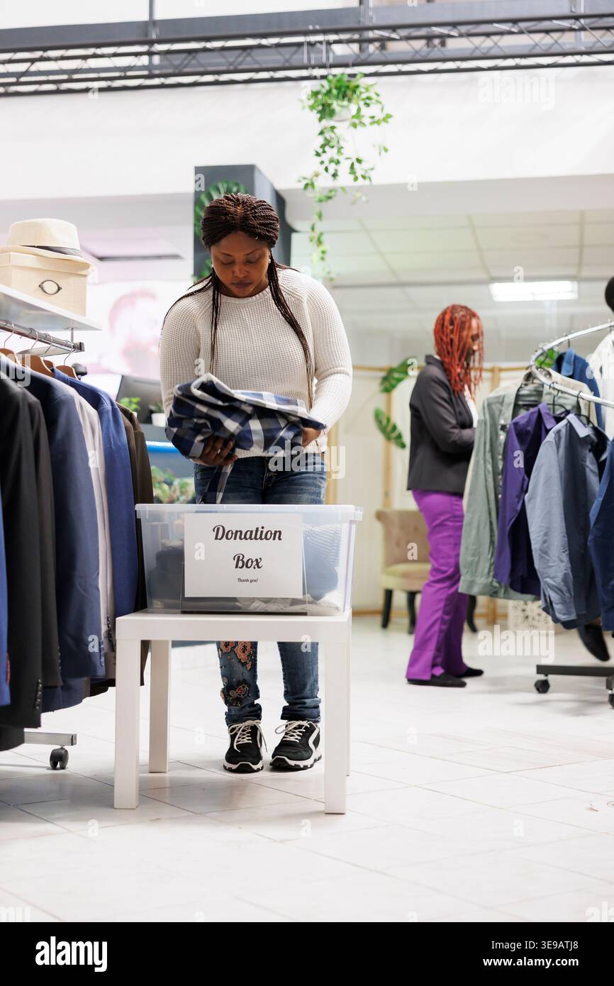 Cliente afroamericano che dona abiti all'organizzazione di welfare nel centro commerciale. Donna che indossa capi di abbigliamento nuovi e di seconda mano in una scatola donazione mentre fa shopping in un negozio alla moda Foto Stock