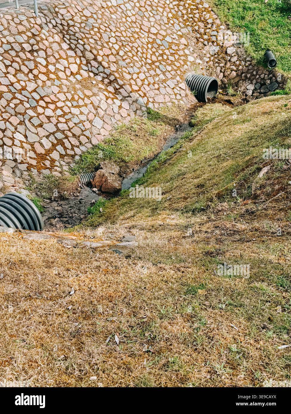 Sistema di drenaggio urbano con tubi scatolari che incanalano le acque piovane attraverso un fossato erboso e un terrapieno rivestito in pietra. Concetto di gestione dell'acqua, erosione Foto Stock
