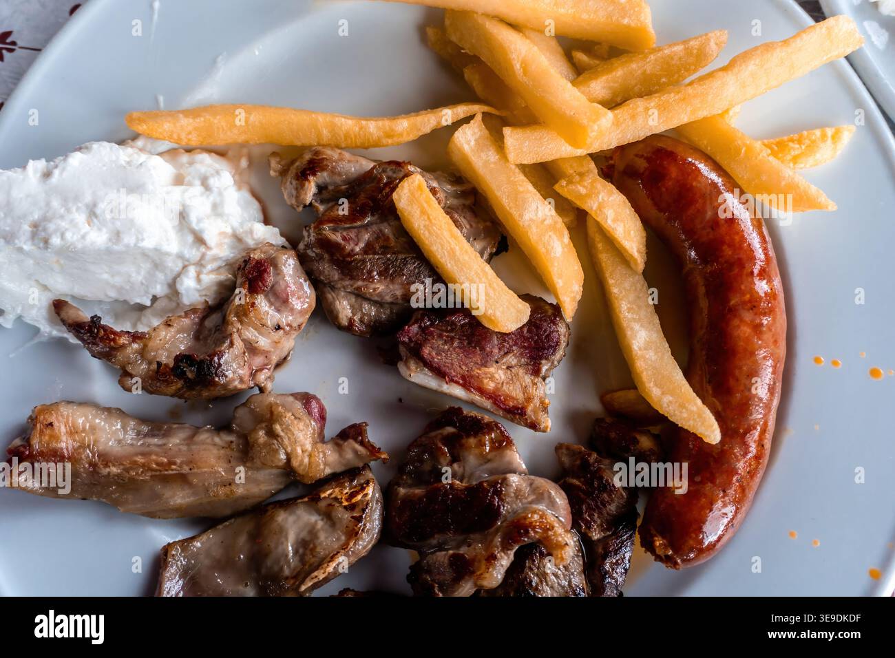 Piatto greco tradizionale con pezzi di maiale alla griglia, salsiccia abbondante, patatine fritte dorate e salsa tzatziki cremosa su un piatto bianco Foto Stock