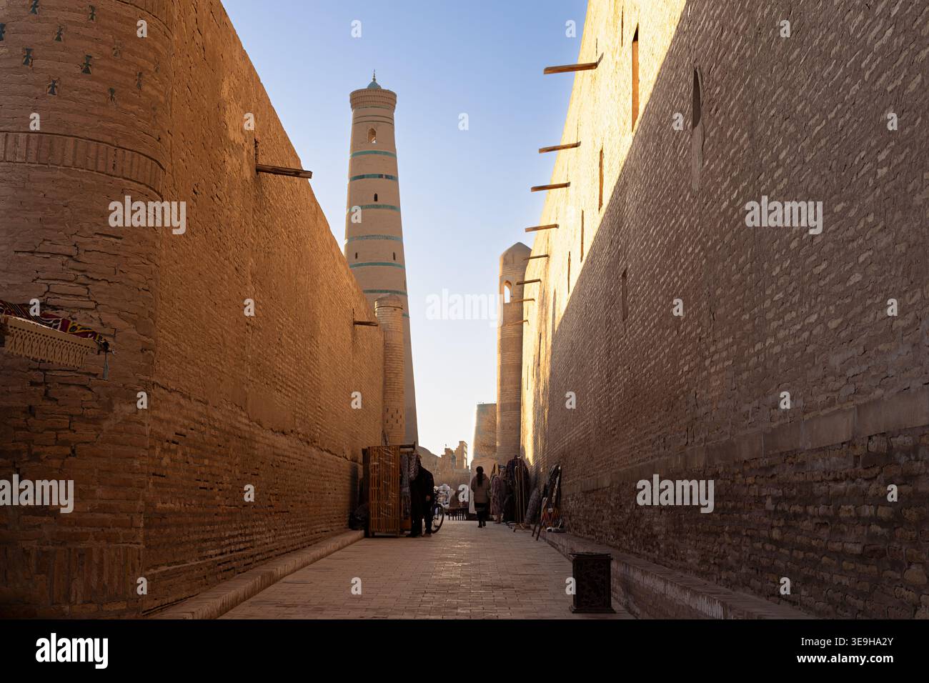 Vicolo stretto nella città vecchia di Khiva di Ichan Kala con minareto che si innalza sopra le mura di mattoni. La calda luce dorata illumina l'antico paesaggio urbano, ideale Foto Stock