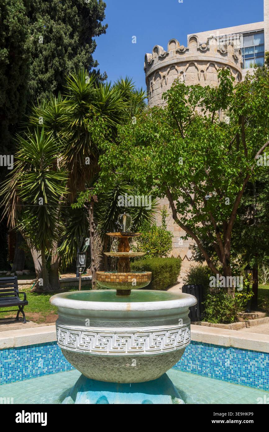 Gerusalemme, Israele, 17 aprile 2026, fontana decorativa in pietra con tre livelli in un soleggiato giardino mediterraneo con palme e una torre del castello sul Foto Stock