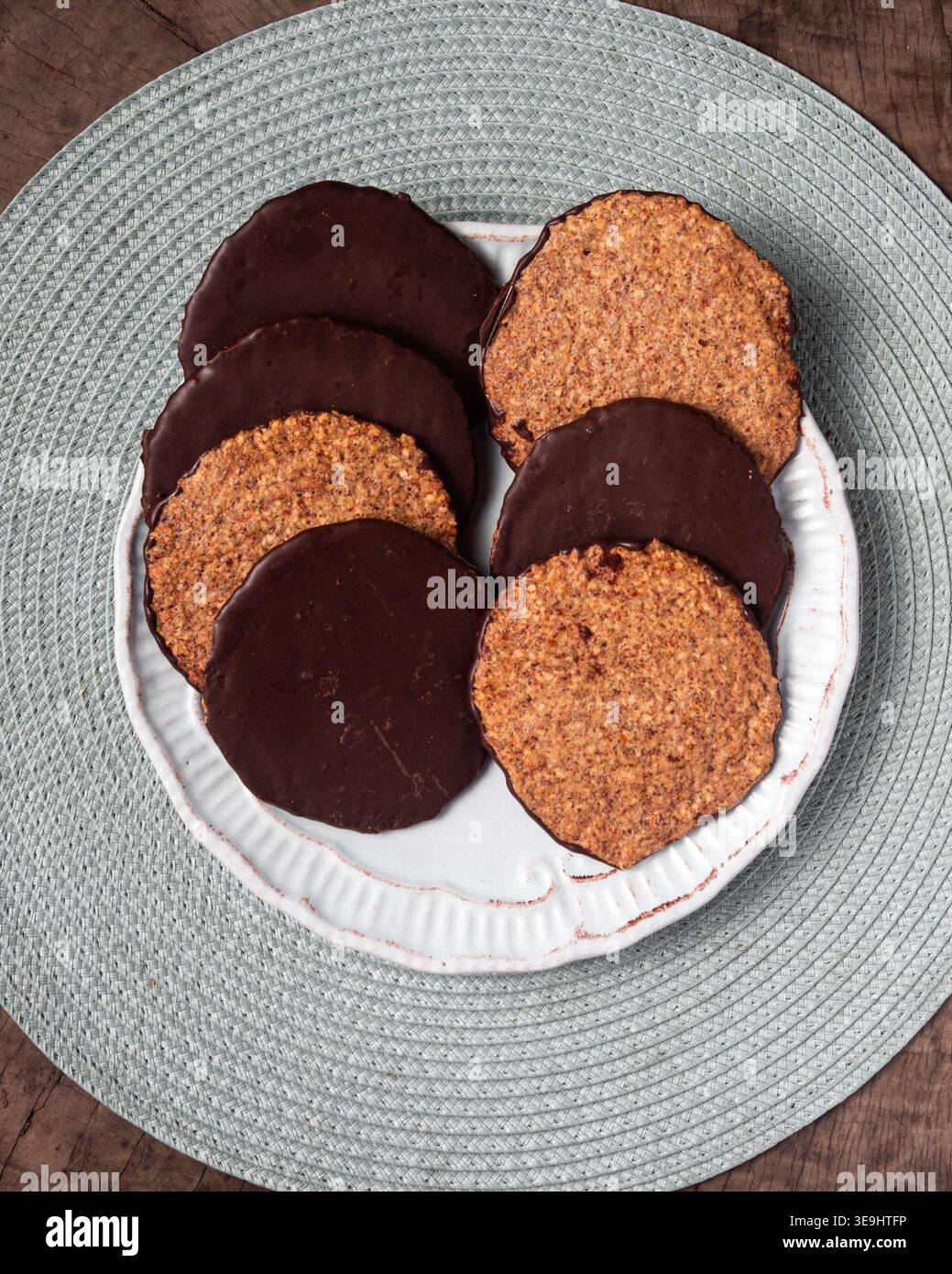 I biscotti rotondi con strati di cioccolato e granulosità si trovano su un piatto bianco. La piastra poggia su una placca grigia testurizzata. La superficie in legno sottostante aggiunge calore, rus Foto Stock