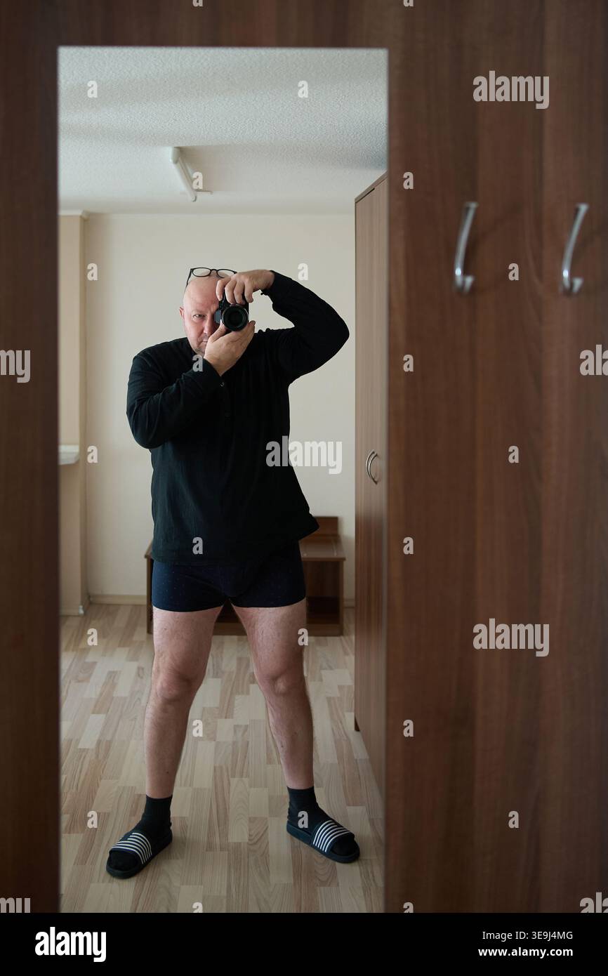 Uomo con maglione nero che scatta un selfie a specchio con una fotocamera in corridoio. Riflesso in un grande specchio su un armadio in legno Foto Stock