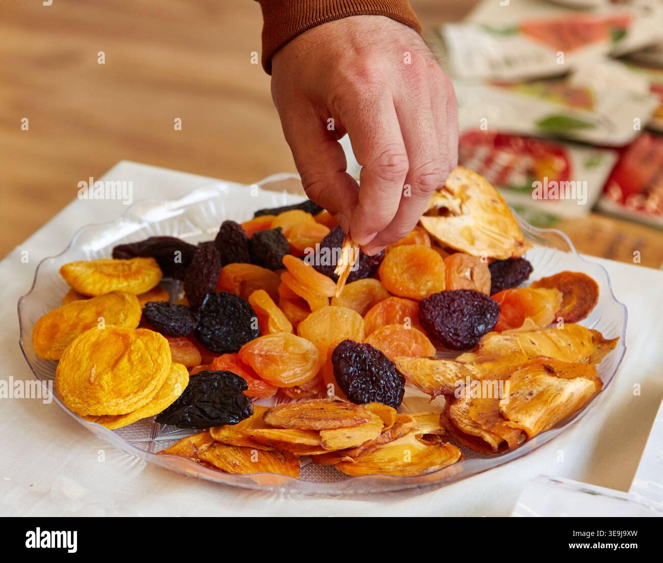 Selezione a mano di vari tipi di frutta secca da un piatto trasparente, tra cui albicocche, prugne e fette di mele, disposte su una tovaglia bianca in un ambiente accogliente Foto Stock