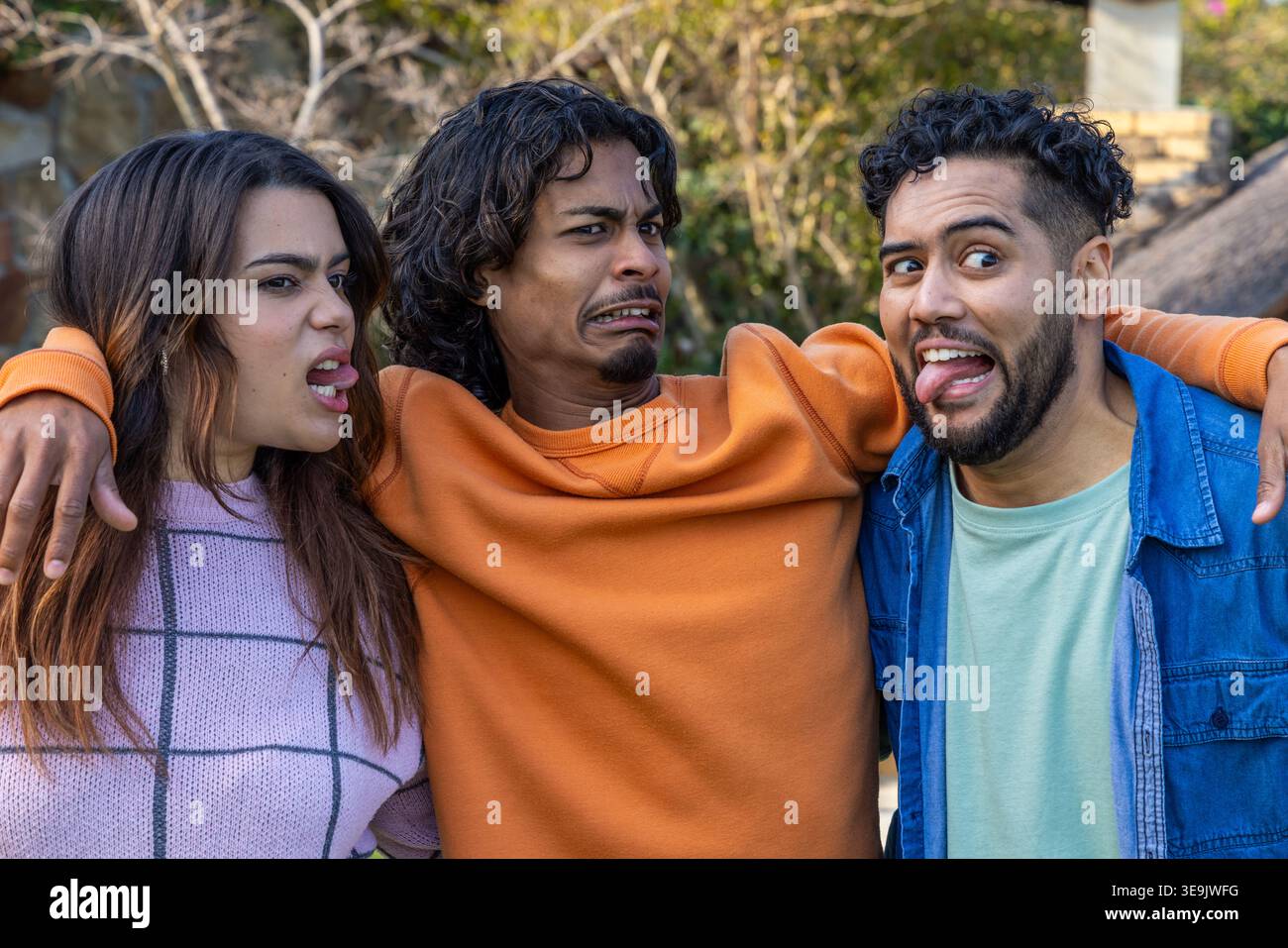 Amici diversi che si fanno i volti in giardino con le braccia legate indossando un maglione in lavanda e un girocollo arancione Foto Stock