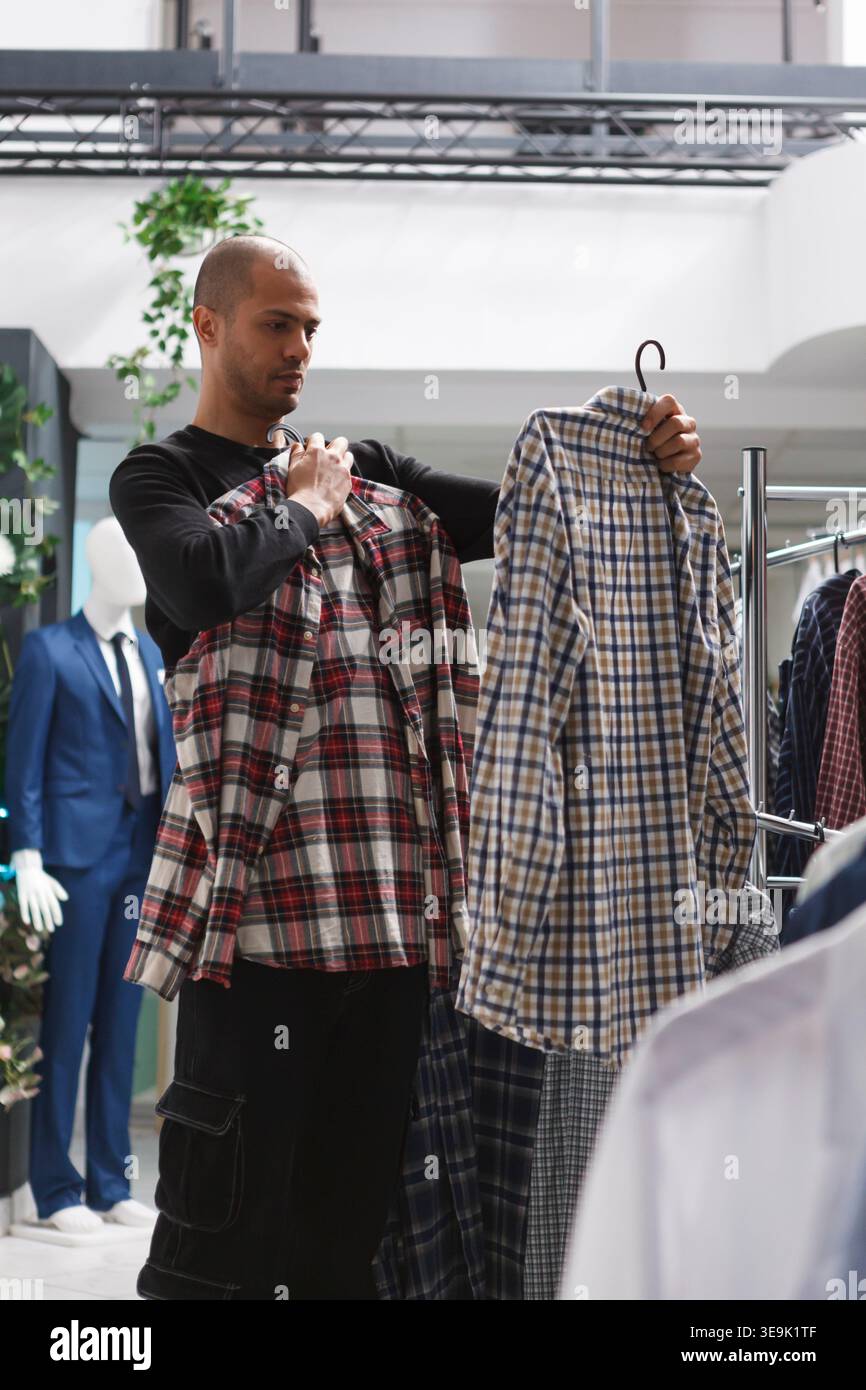 Uomo arabo premuroso che esamina due magliette prima di prendere una decisione di acquisto in un negozio di abbigliamento. Centro commerciale boutique che controlla lo stile e le dimensioni dell'abbigliamento mentre scegli l'abbigliamento Foto Stock