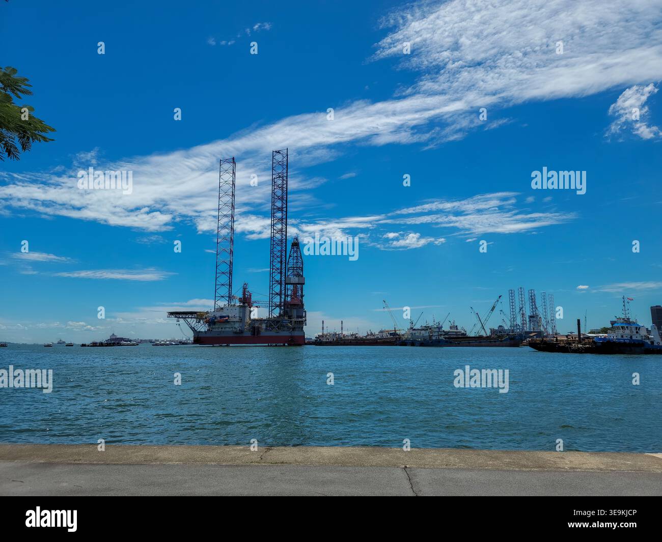 Il grande carro di perforazione offshore galleggia in acque blu calme. Il cielo è chiaro con nuvole bianche sparse sopra. Navi e barche distanti costellano l'orizzonte del porto Foto Stock
