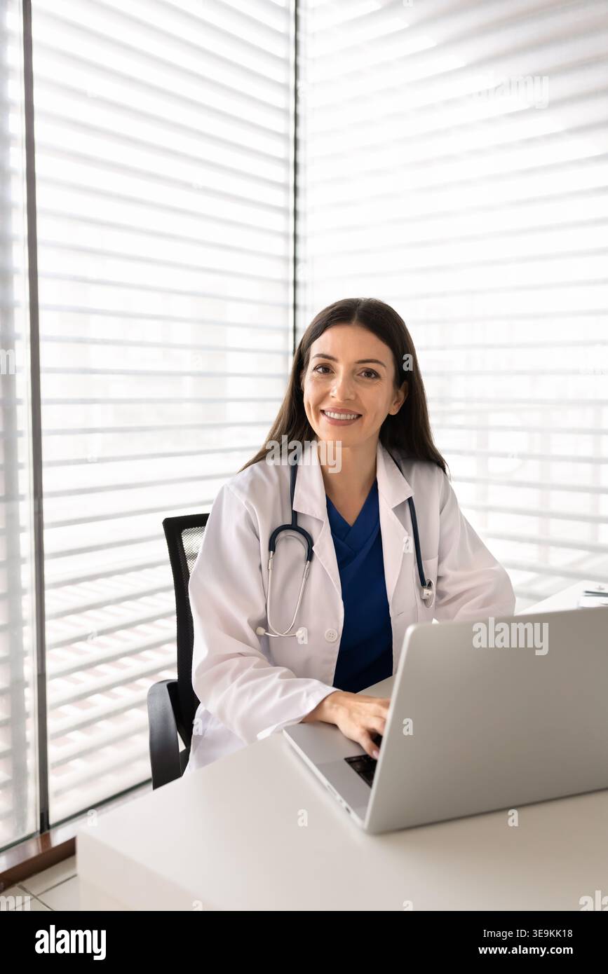 Medico che guarda la macchina fotografica, seduto alla scrivania con il computer portatile Foto Stock