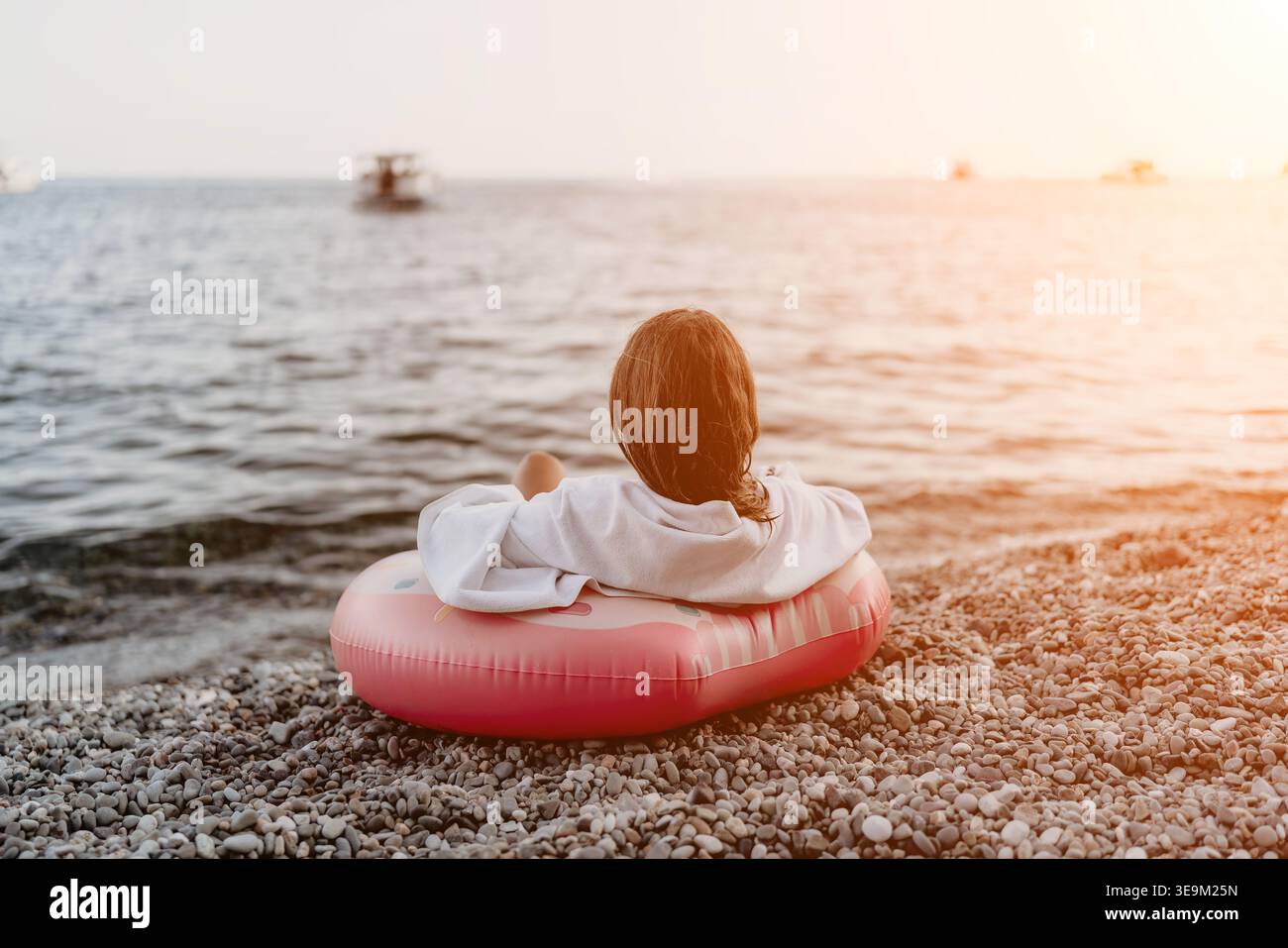 Donna, spiaggia, tramonto. Vista posteriore di una donna che si rilassa su un anello gonfiabile rosa in una spiaggia di ciottoli durante l'ora d'oro con spazio per fotocopie. Foto Stock