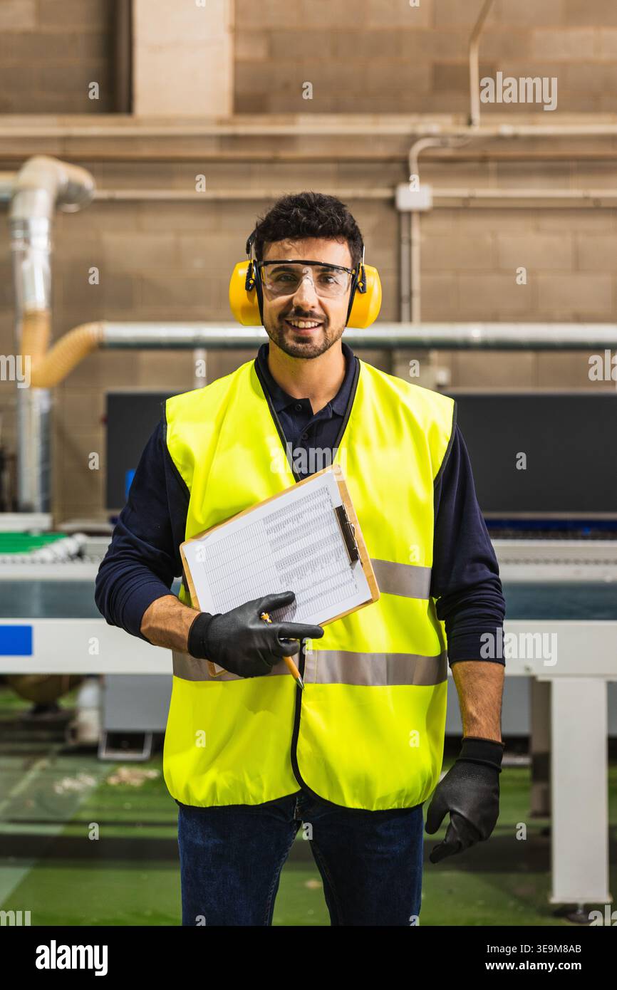 Lavoratore sorridente che indossa giubbotto di sicurezza e cuffie in fabbrica Foto Stock