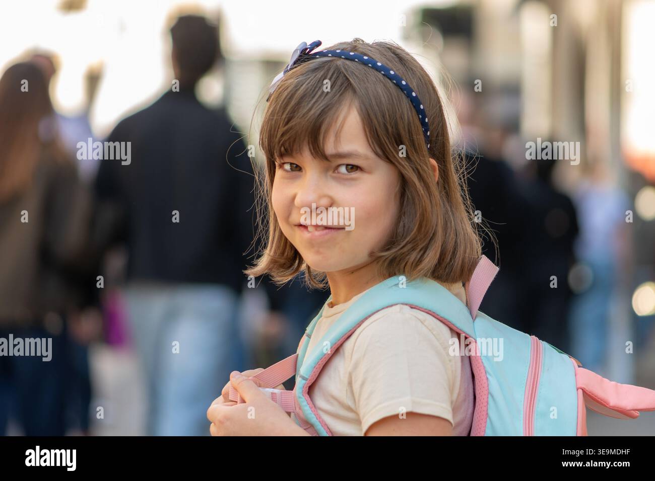 Ragazza di 7 anni che cammina con lo zaino guardando indietro nella scena urbana. Foto Stock