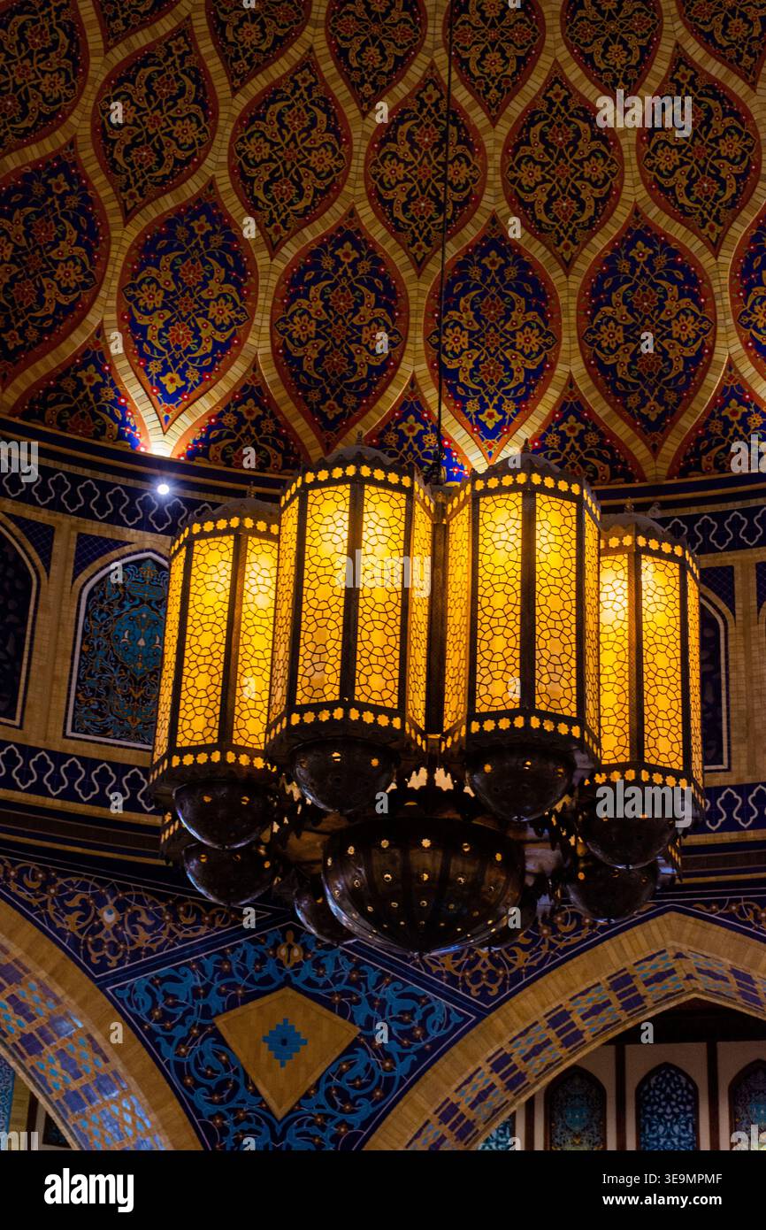 Lampadario decorato e luminoso in stile arabo appeso sotto un soffitto geometrico intricatamente decorato che presenta arte islamica e piastrelle. Foto Stock