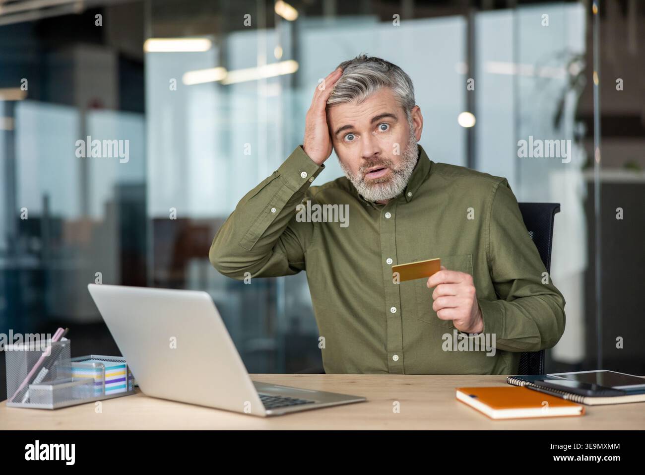 Uomo maturo con un'espressione sorpresa e scioccata che tocca la testa, si trova in una situazione finanziaria inaspettata mentre tiene in mano una carta di credito e guarda un notebook in ufficio Foto Stock
