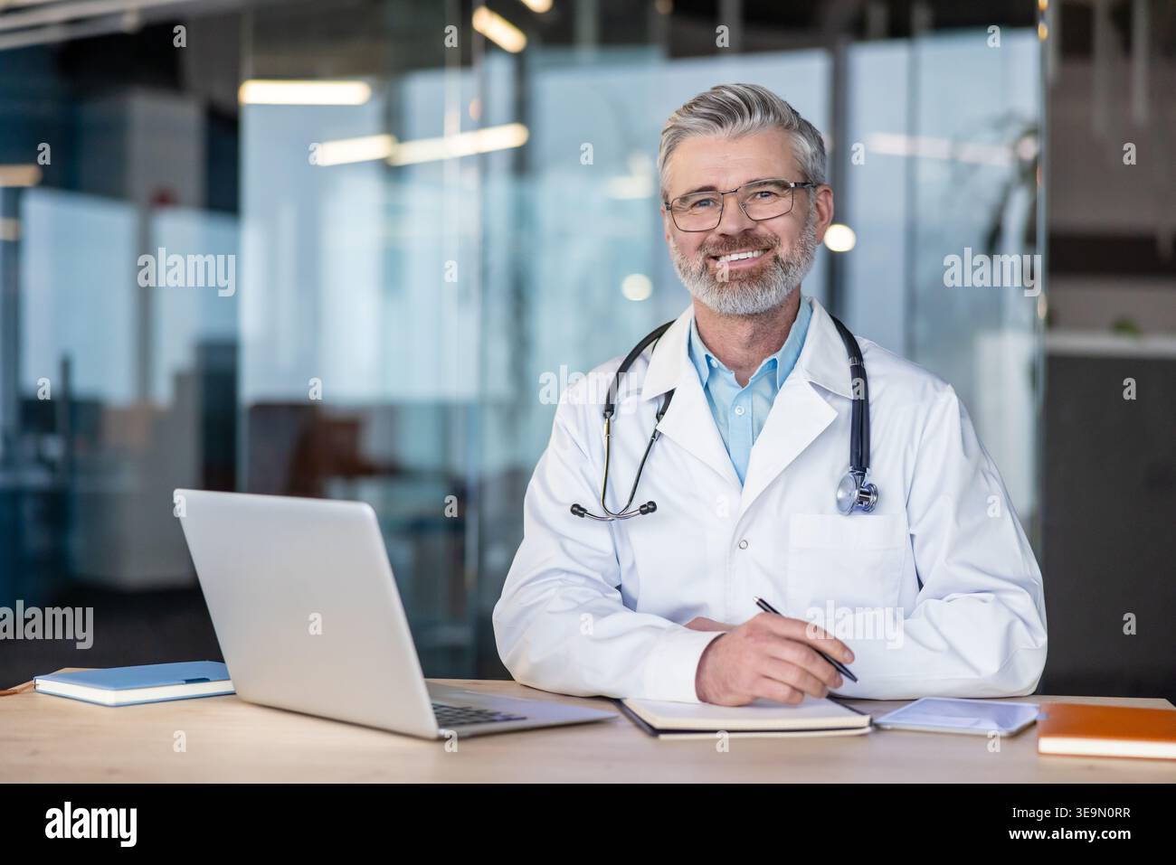 Medico di sesso maschile esperto che indossa un camice da laboratorio e uno stetoscopio, sorridendo mentre è seduto alla sua scrivania con un portatile e un notebook, che rappresenta l'assistenza sanitaria, il benessere e il servizio medico professionale Foto Stock