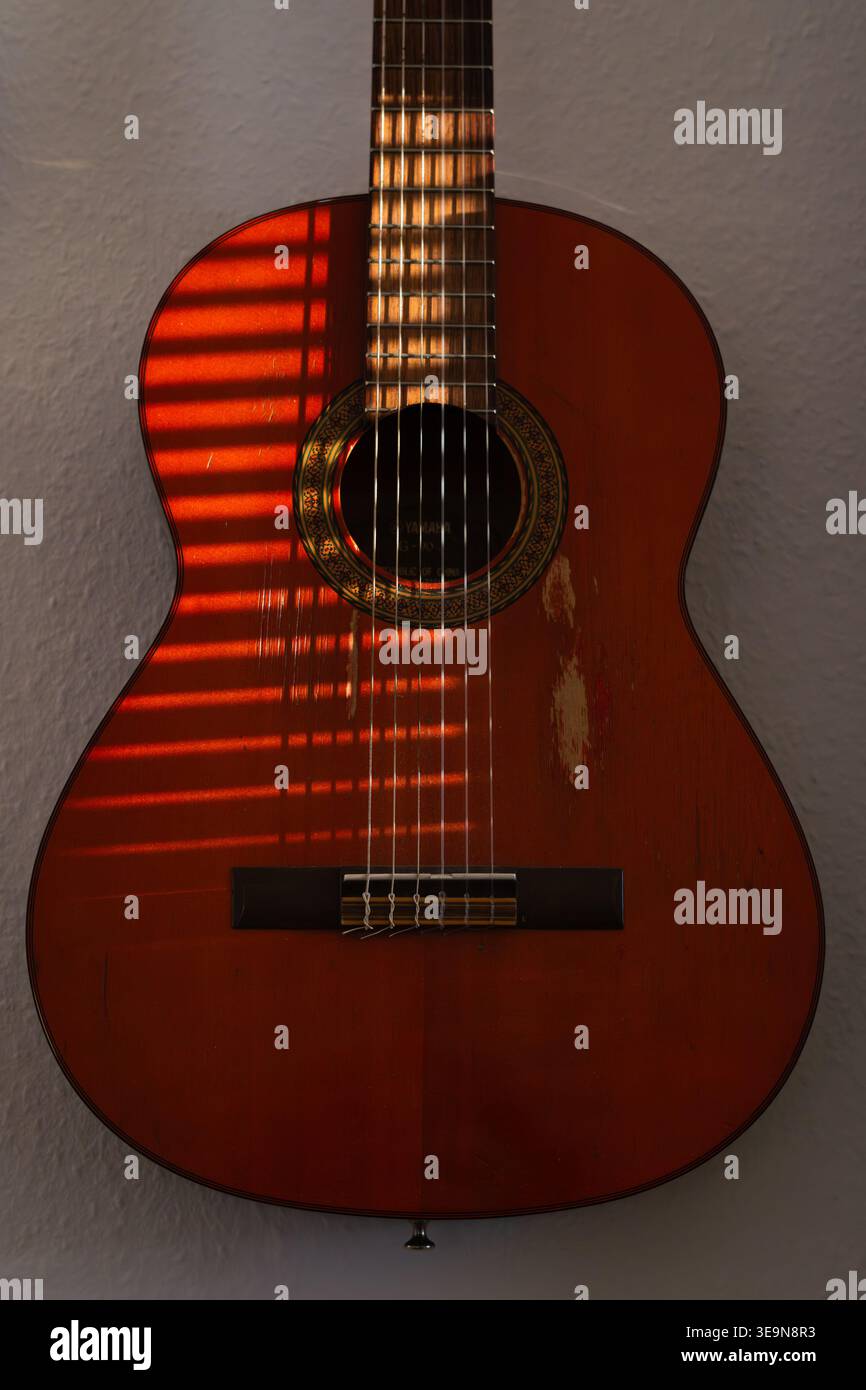 Una chitarra molto amata e suonata appesa a un muro. La luce dalla finestra proietta ombre a righe sul corpo e sul collo della chitarra. Foto Stock