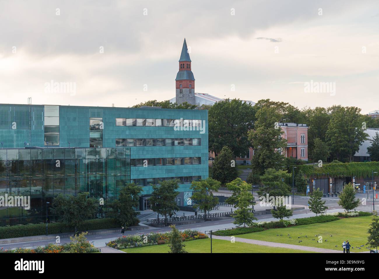 HELSINKI, FINLANDIA - 11 AGOSTO 2025: Edificio per uffici in vetro turchese con una torre in mattoni rossi che si innalza sopra alberi verdi in estate. Foto Stock