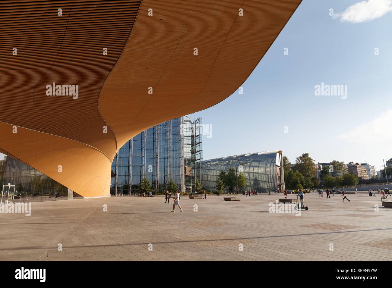 HELSINKI, FINLANDIA - 11 AGOSTO 2025: Ampia piazza pubblica con un baldacchino dorato curvo e un edificio a torre di vetro sotto un cielo estivo. Foto Stock