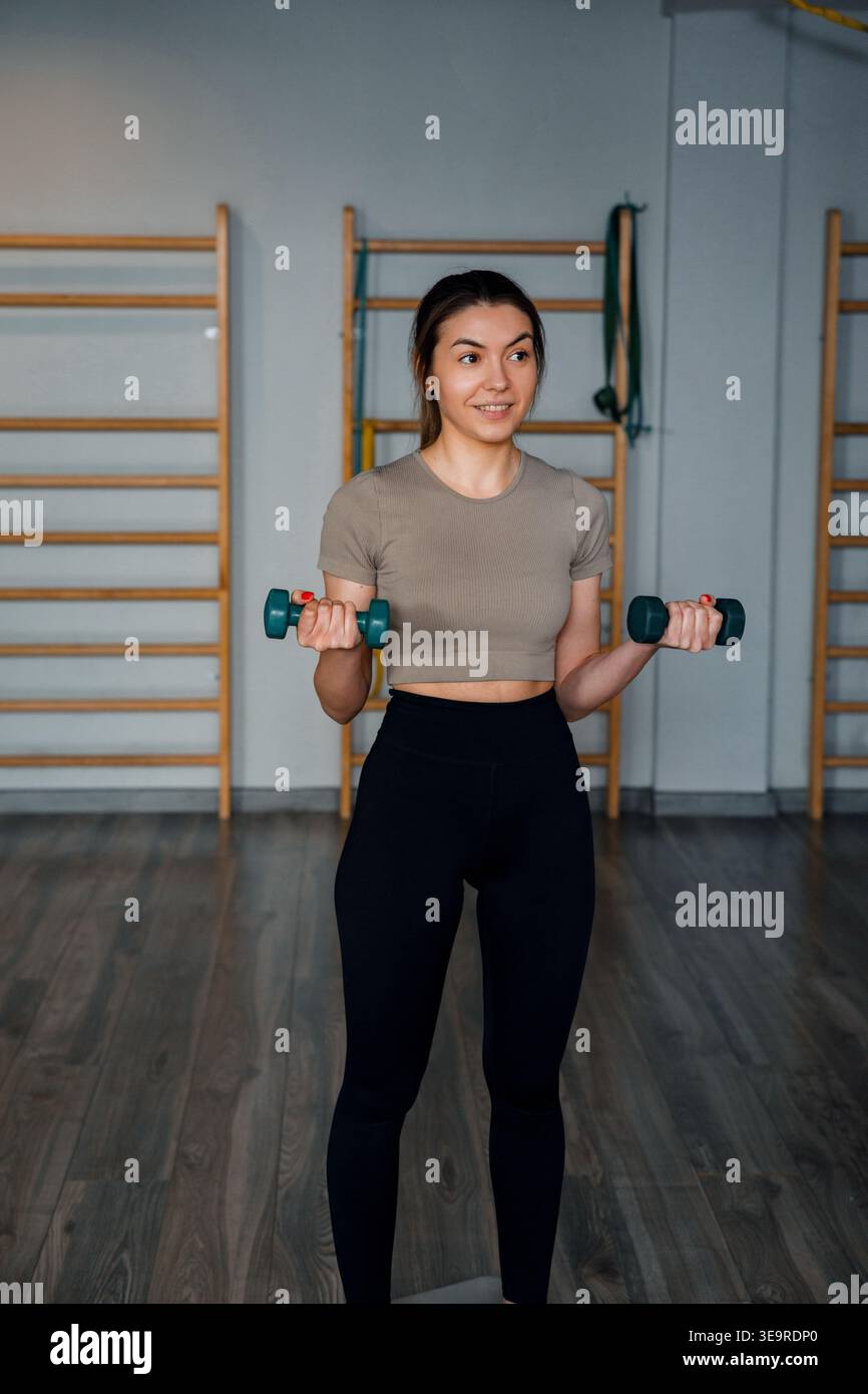 Concetto di fitness femminile all'interno della palestra. Giovane donna che tiene i manubri. Modello femminile per l'allenamento della parte superiore del corpo. Sessione di allenamento attiva al chiuso. Healt Foto Stock