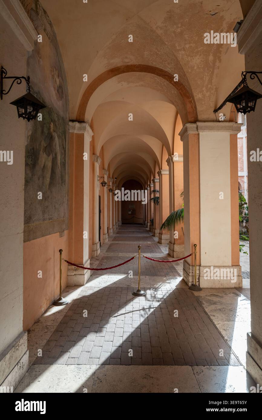 Roma, Italia - 28 agosto 2025: Corridoio portico a volta illuminato dal sole con archi, lanterne sospese e barriera di corda presso la Basilica dei Santi Cosma e Damia Foto Stock