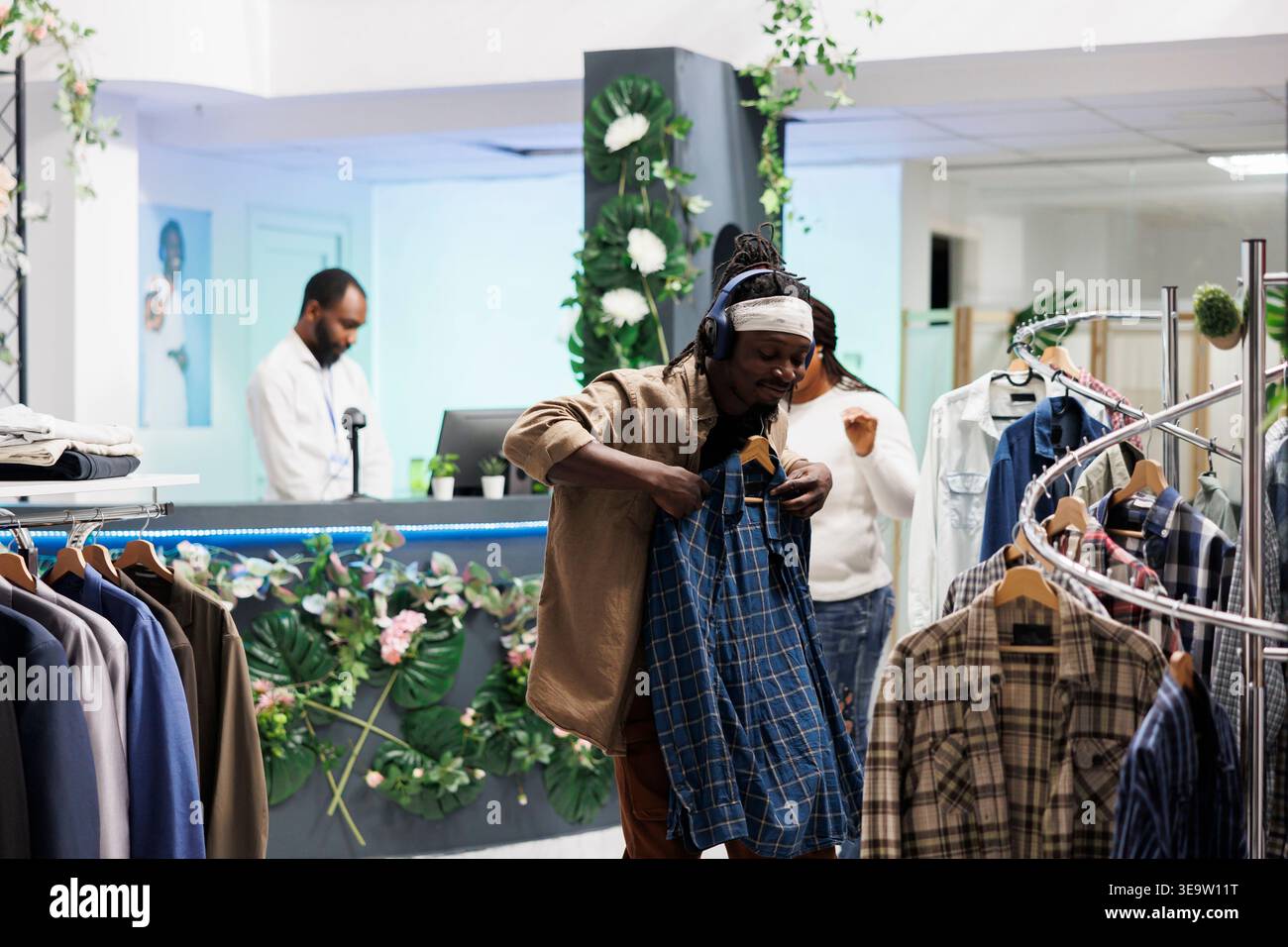 Allegro uomo afroamericano che acquista abbigliamento mentre si ascolta la musica in cuffia. Cliente di un negozio di abbigliamento con cuffie che tiene la camicia sull'appendiabiti mentre sceglie un abbigliamento in boutique Foto Stock