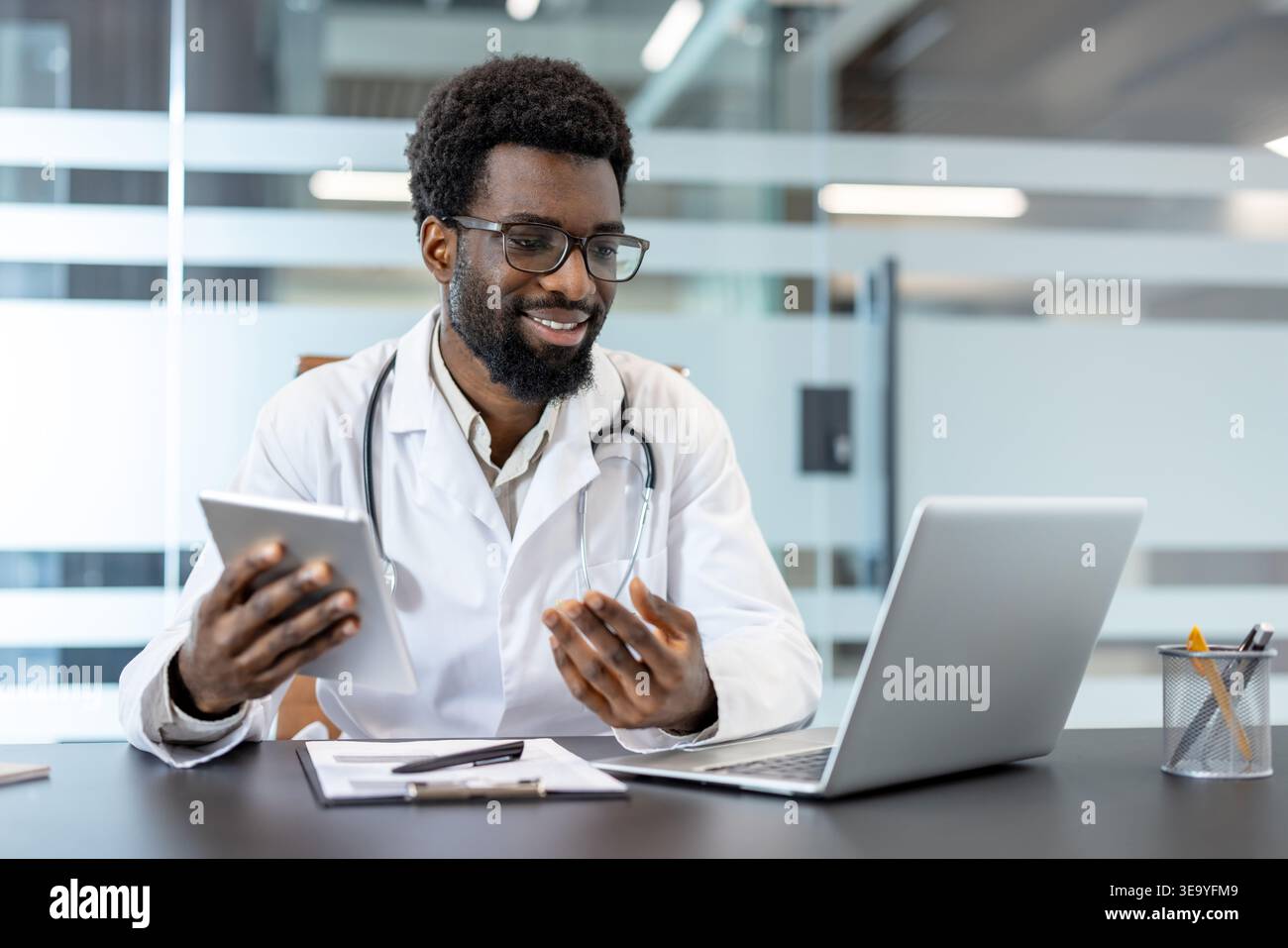 Medico afroamericano che indossa un camice da laboratorio e uno stetoscopio. Sorridere mentre si utilizza un tablet digitale e un notebook alla scrivania. Fornire consulenza medica virtuale e adottare la moderna tecnologia sanitaria Foto Stock