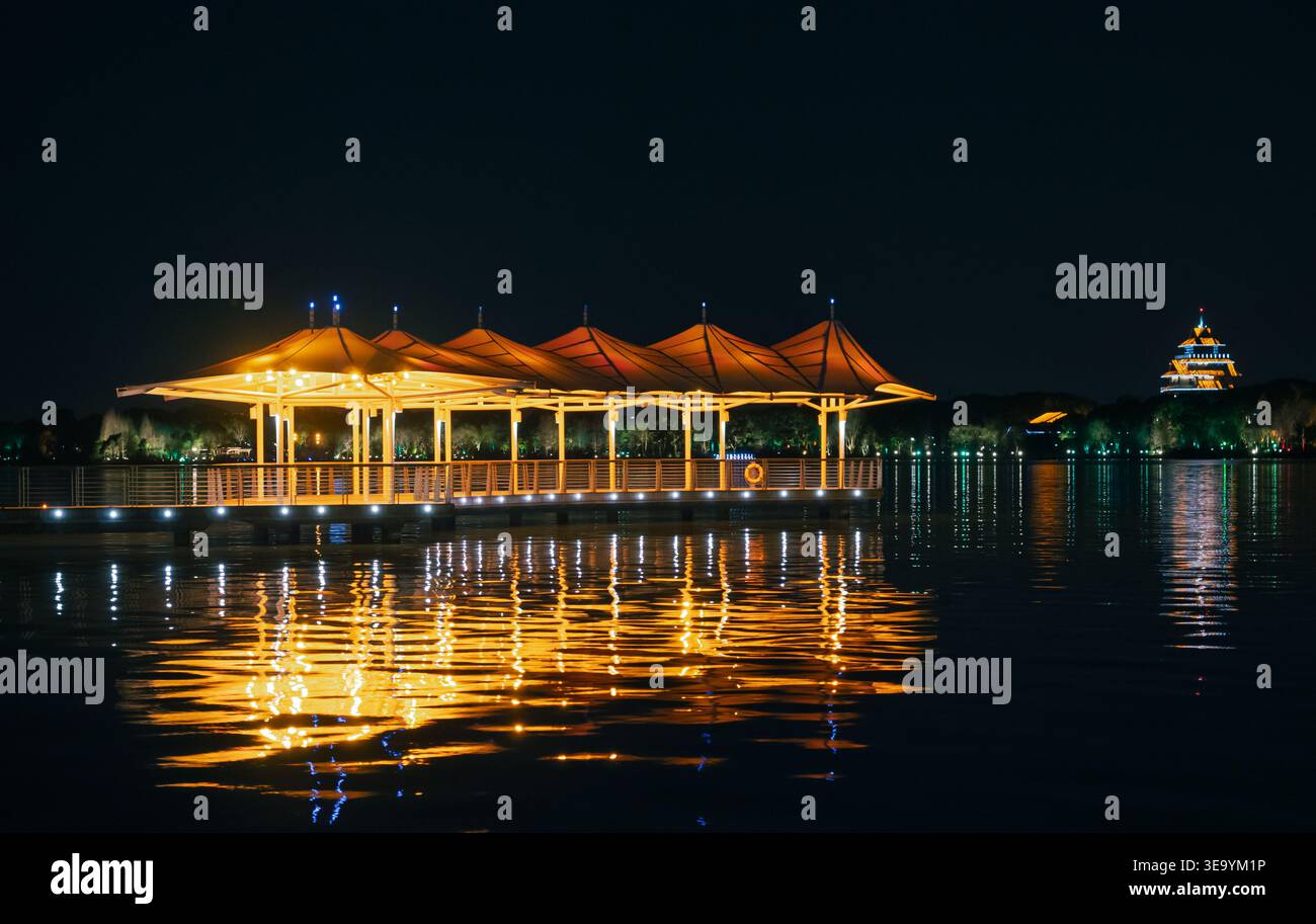 Padiglioni dal tetto dorato illuminano un molo panoramico del lago, che si riflette sulle acque tranquille di notte, con una lontana pagoda che si illumina Foto Stock