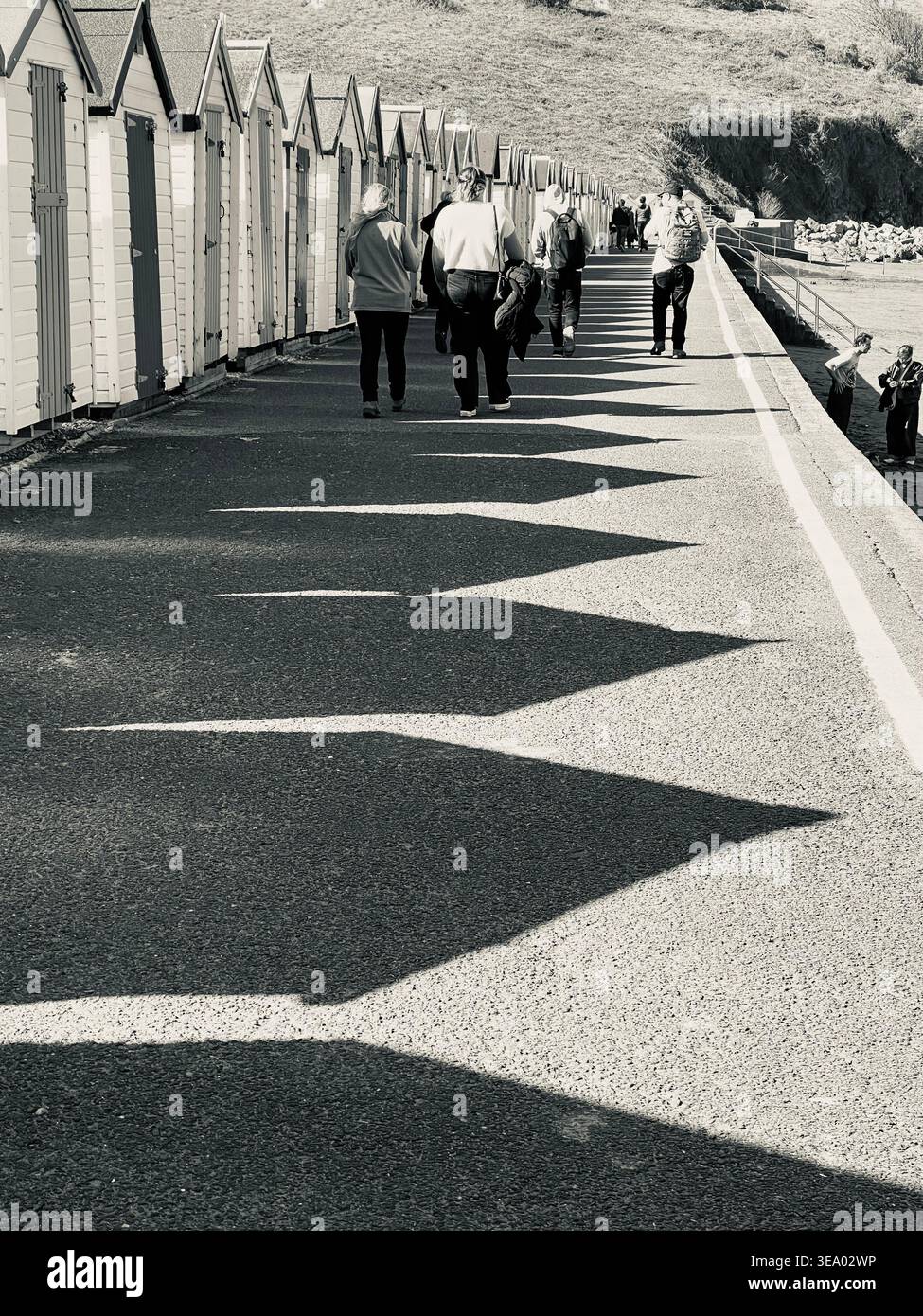 Immagine in bianco e nero di pedoni che camminano lungo la passeggiata di Broadsands, passando per capanne sulla spiaggia che gettano un'ombra profonda sul marciapiede, Devon, Regno Unito Foto Stock