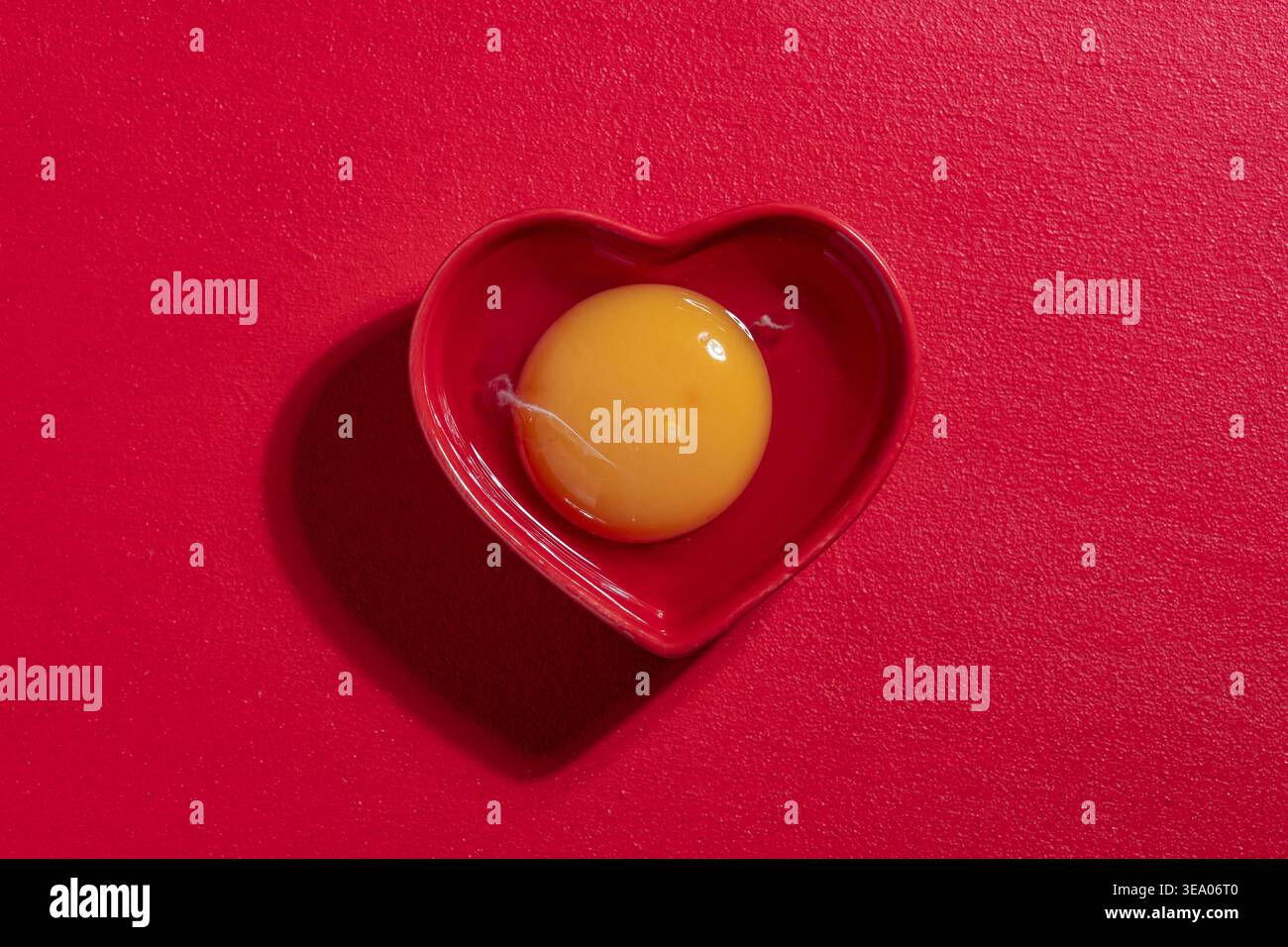 Tuorlo d'uovo di pollo crudo in un recipiente di ceramica a forma di cuore rosso su fondo rosso testurizzato. Fotografia gastronomica minimalista dall'alto per San Valentino o per la guarigione Foto Stock