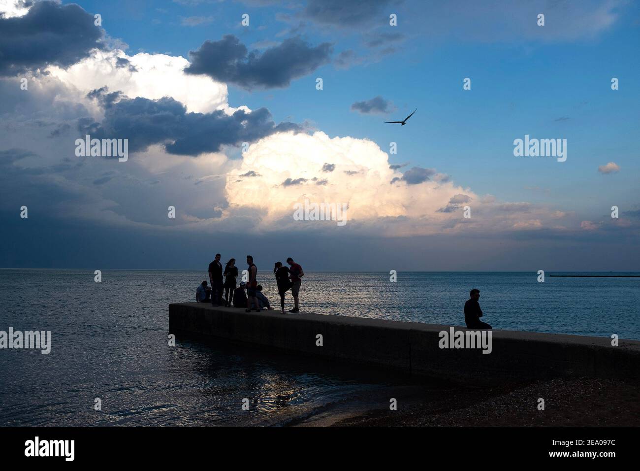 Le persone stanno su un molo mentre una persona siede da sola sul bordo dell'acqua sotto un cielo spettacolare con nuvole e un uccello che vola sopra la testa. Foto Stock