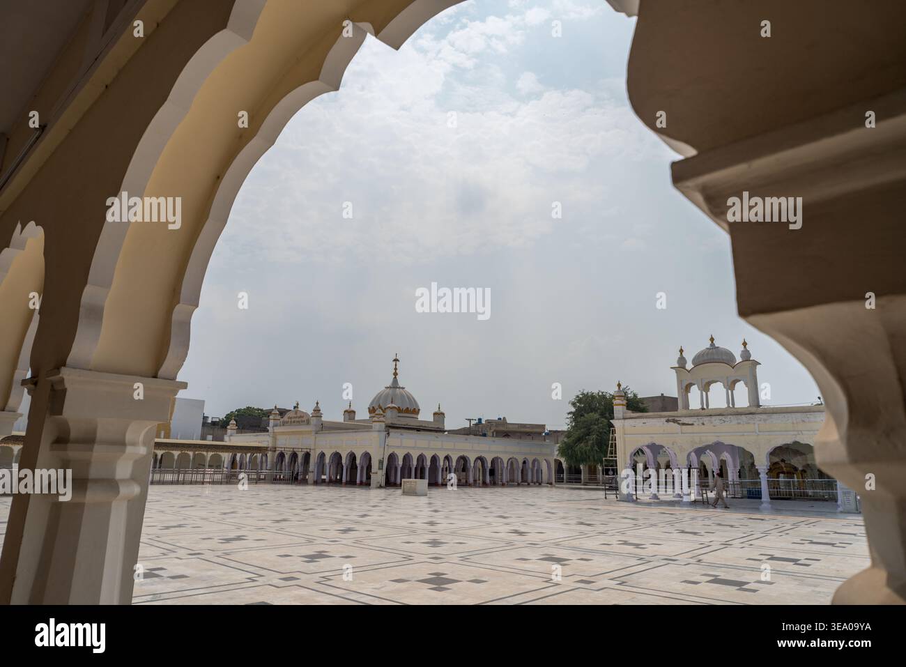 Vista di un tranquillo cortile con colonnati ad arco color crema che incorniciano un edificio lontano e ornato sotto un cielo morbido, Nankana Sahib, Punjab, Pakistan. Foto Stock