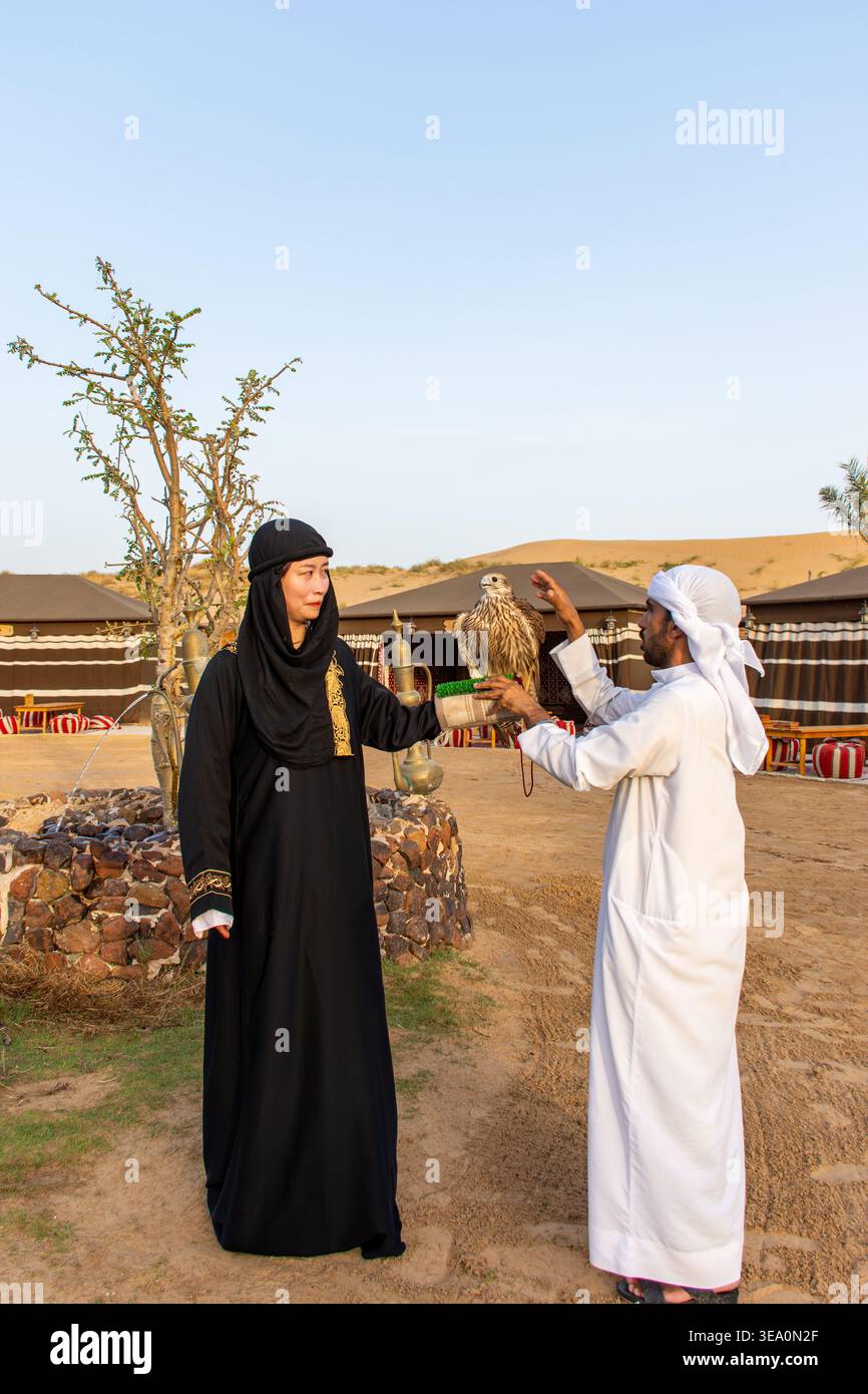 Donna turistica in una tradizionale abaya nera che tiene in mano un falco mentre un uomo arabo locale in una kandura bianca la assiste in un campo safari nel deserto a Dubai. Foto Stock