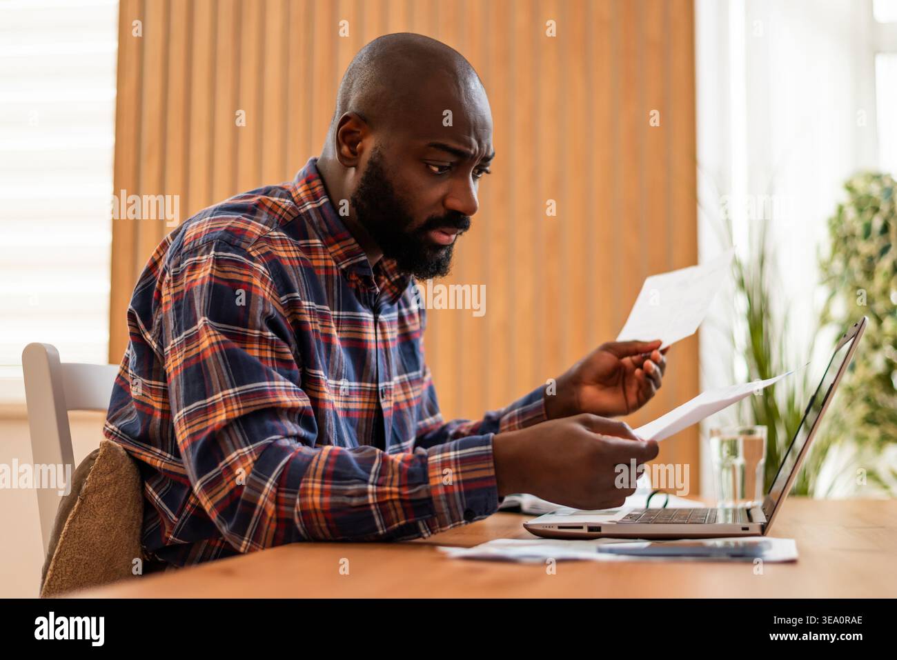 L'uomo analizza le bollette finanziarie mentre è seduto a un notebook, sembrando sopraffatto dalla sua situazione finanziaria. Foto Stock