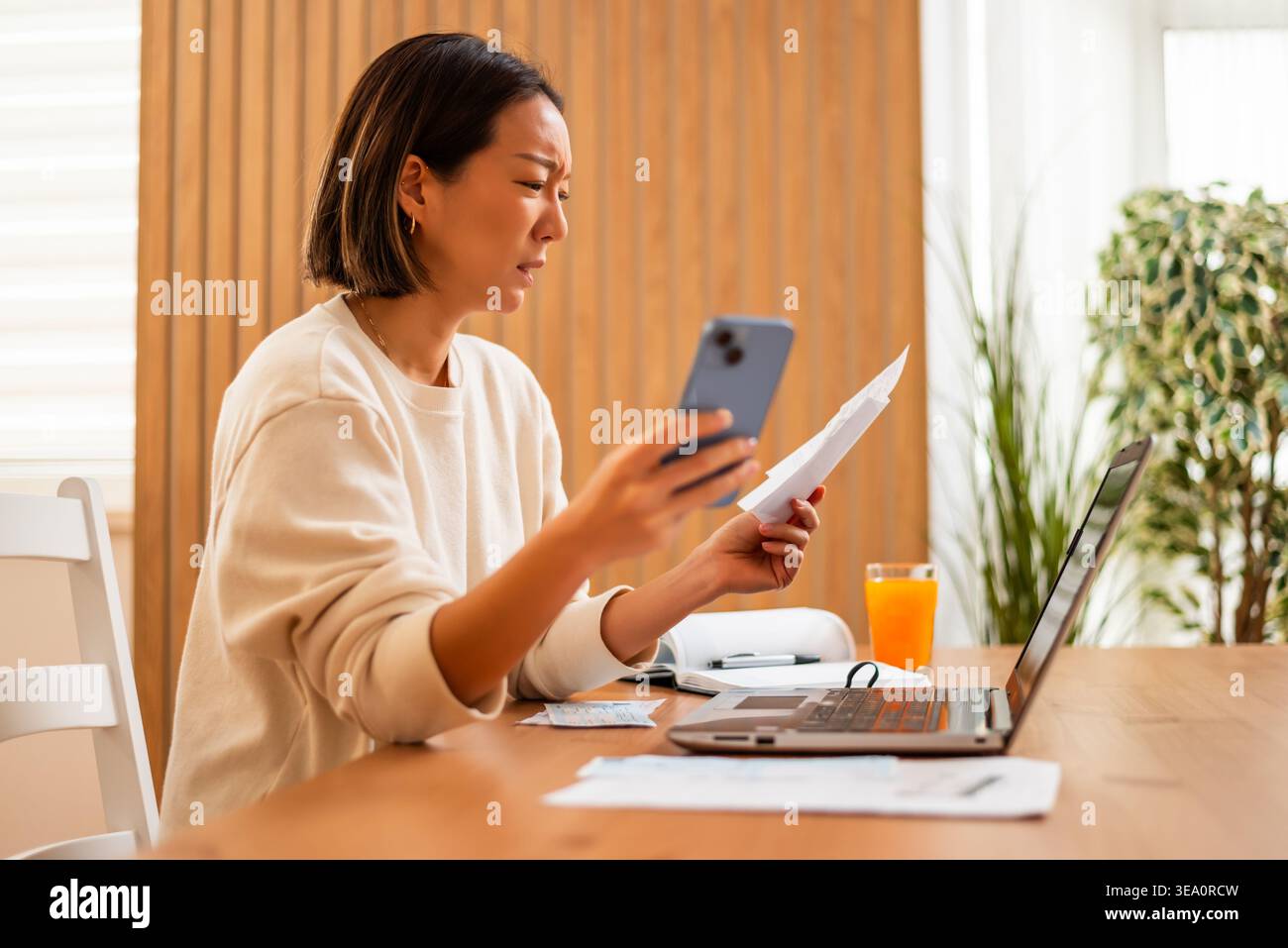 Una donna analizza i documenti e la sua carta di credito mentre è seduta davanti a un portatile, apparendo sopraffatta dalla sua situazione finanziaria. Foto Stock