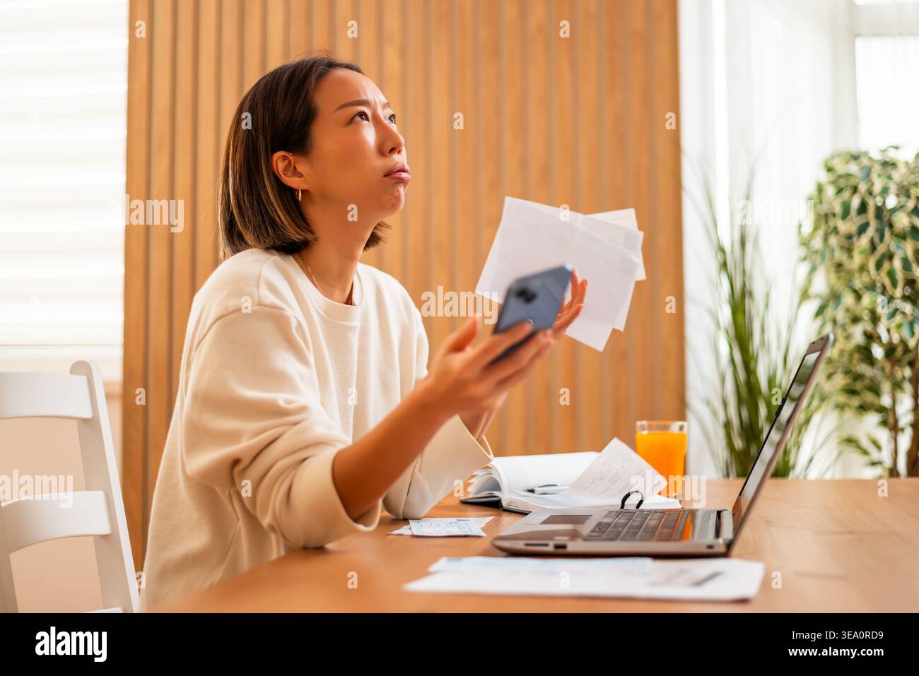 Una donna analizza i documenti e la sua carta di credito mentre è seduta davanti a un portatile, apparendo sopraffatta dalla sua situazione finanziaria. Foto Stock