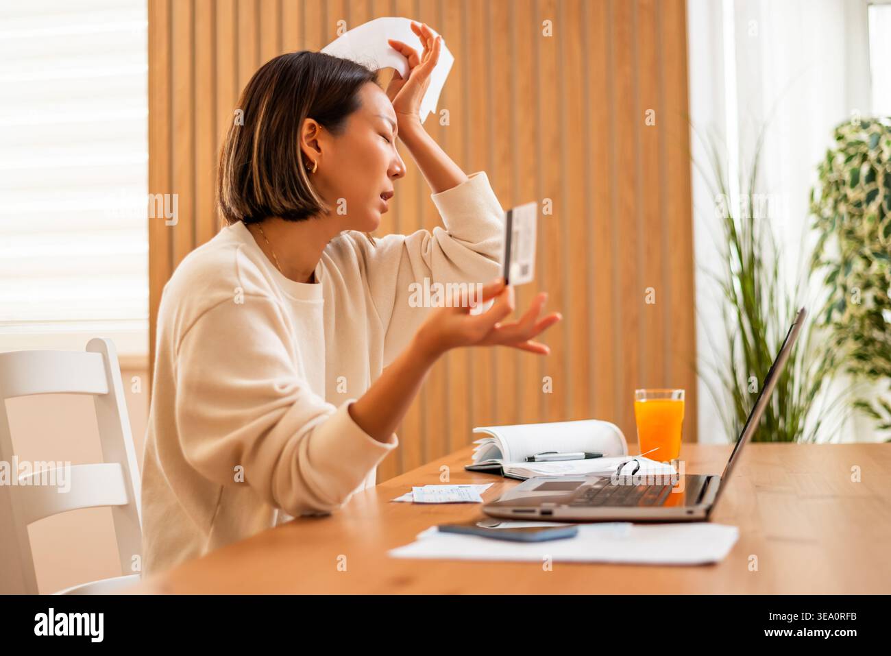 Una donna analizza i documenti e la sua carta di credito mentre è seduta davanti a un portatile, apparendo sopraffatta dalla sua situazione finanziaria. Foto Stock