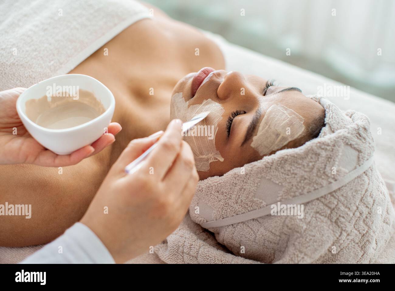 Estetista applicando maschera di argilla bianca con spatola su giovane donna nera Foto Stock