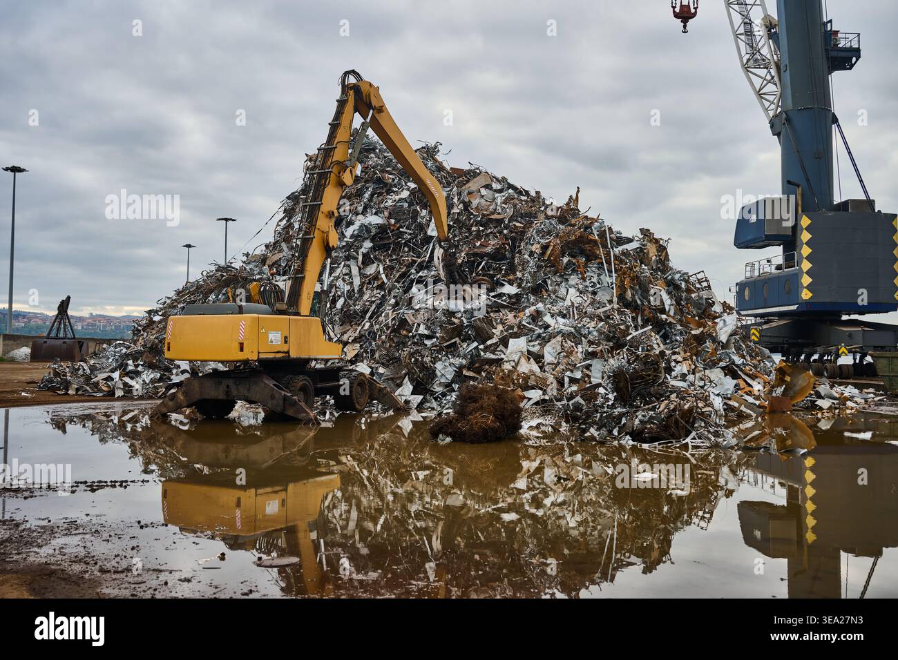 Sito industriale con una grande pila di rottami metallici assortiti, un escavatore e una gru portuale che si prepara per il processo di riciclaggio Foto Stock