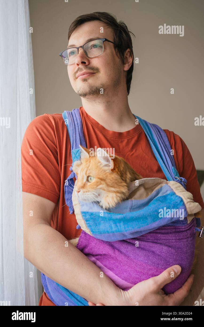 Uomo che indossa una t-shirt arancione e occhiali che tiene in mano il suo amato gatto allo zenzero arancione comodamente accoccolato in un elegante portabicchieri in tessuto all'interno di una casa, mostrando il forte legame animale-umano domestico Foto Stock