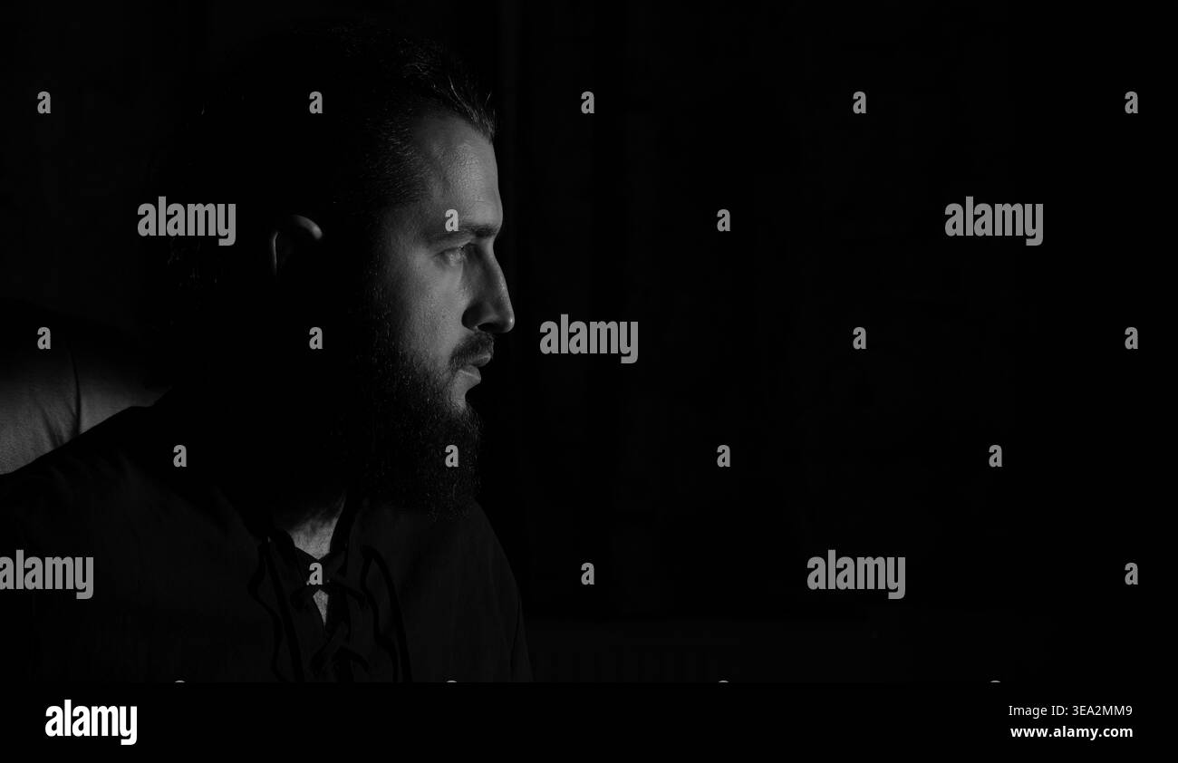 Un uomo con la barba folta viene catturato in un profilo bianco e nero, il suo volto è illuminato da una luce spettacolare su uno sfondo scuro Foto Stock