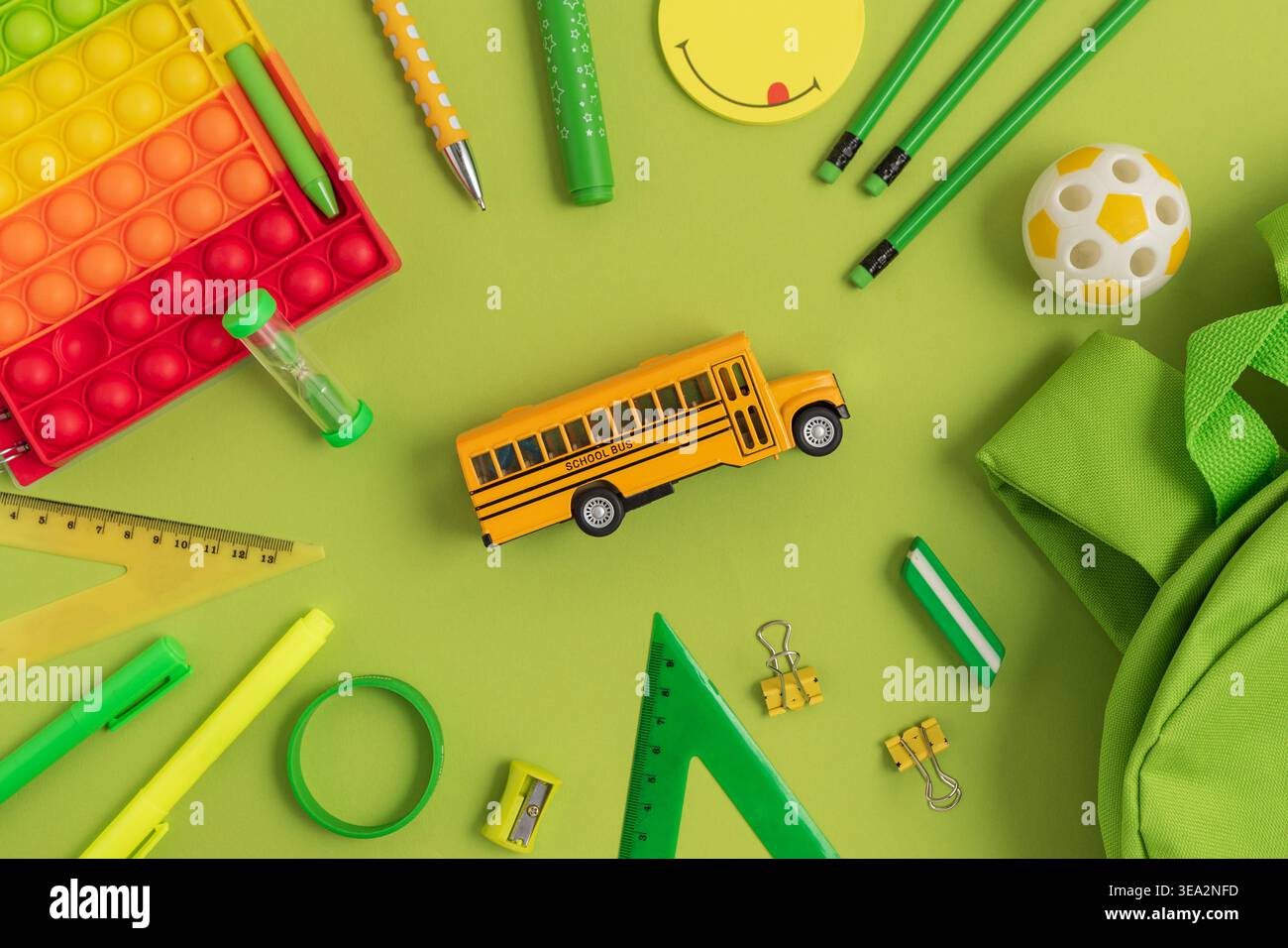 Divertente composizione piatta con scuolabus con materiale scolastico verde e giallo, tra cui blocco note, penne, matite, zaino, righello, clip, t Foto Stock