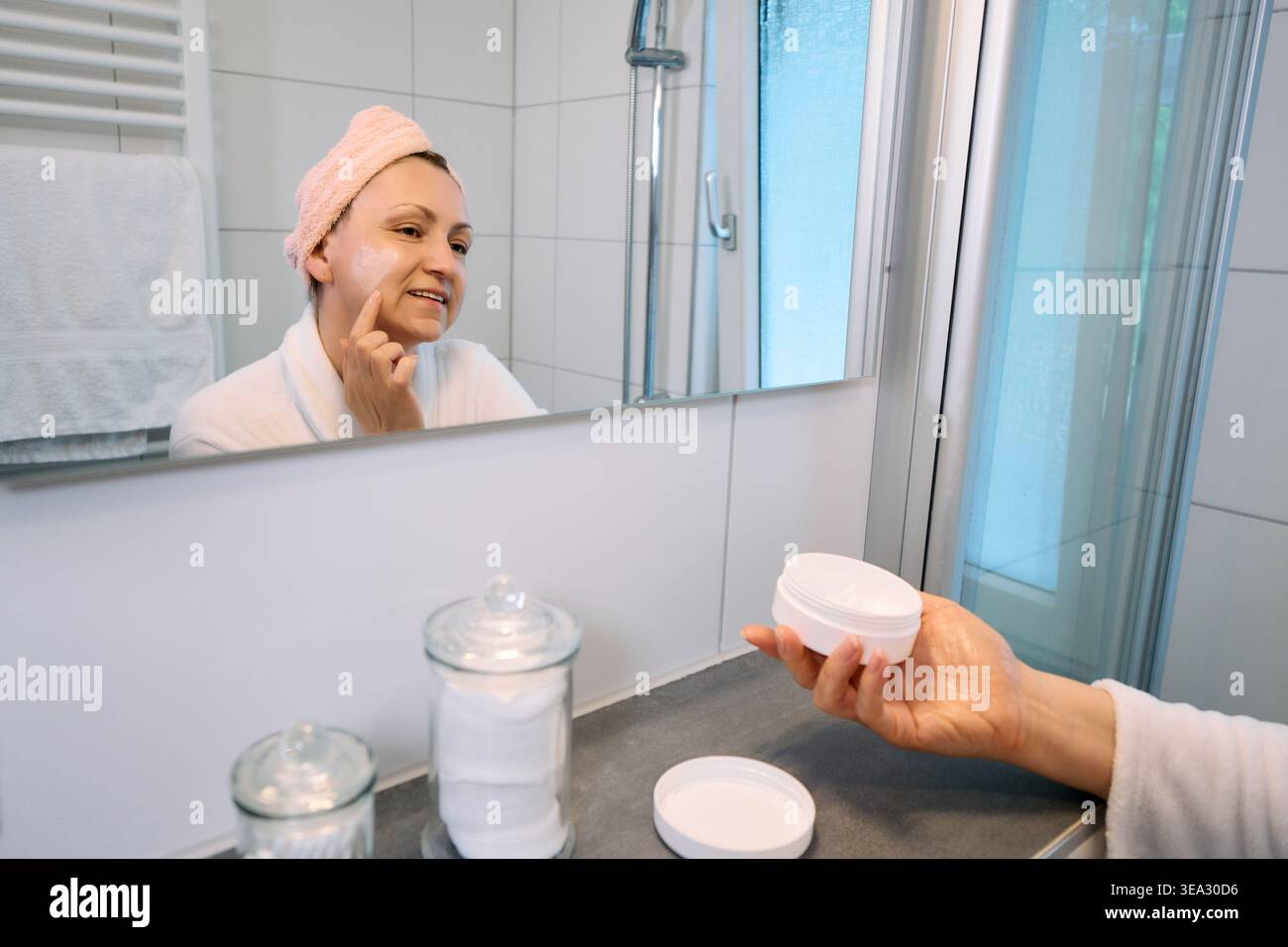 Donna di mezza età che applica crema viso, guarda nello specchio in un bagno luminoso, regge un vaso cosmetico bianco, cura della pelle e concetto di invecchiamento. Foto Stock