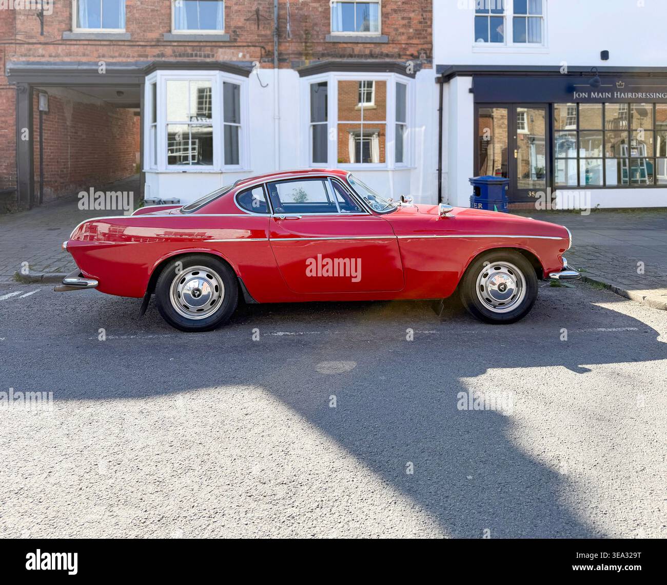 Vista profilo di una Volvo P1800 Classic coupé rossa del 1969 parcheggiata in una strada di fronte a edifici tradizionali in mattoni e parrucchiere a Eccleshall, Inghilterra - Immagine stock catturata con smartphone