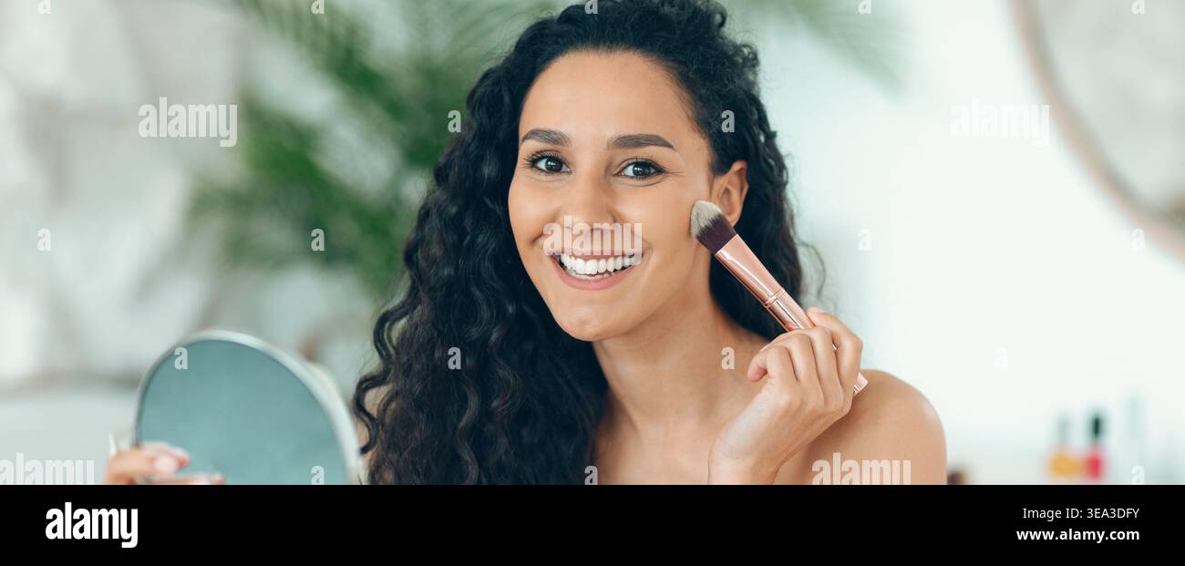 Donna che applica il trucco in uno spazio interno luminoso mentre guarda allo specchio e sorride Foto Stock